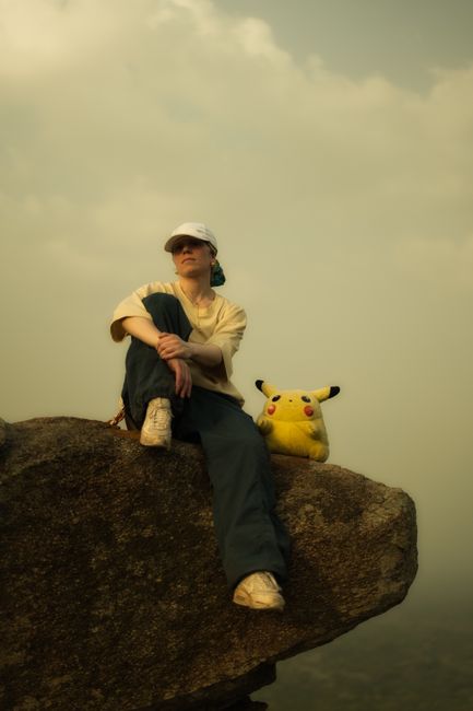 Person in cap and casual attire sitting with Pikachu plush on a mountain rock under cloudy sky.