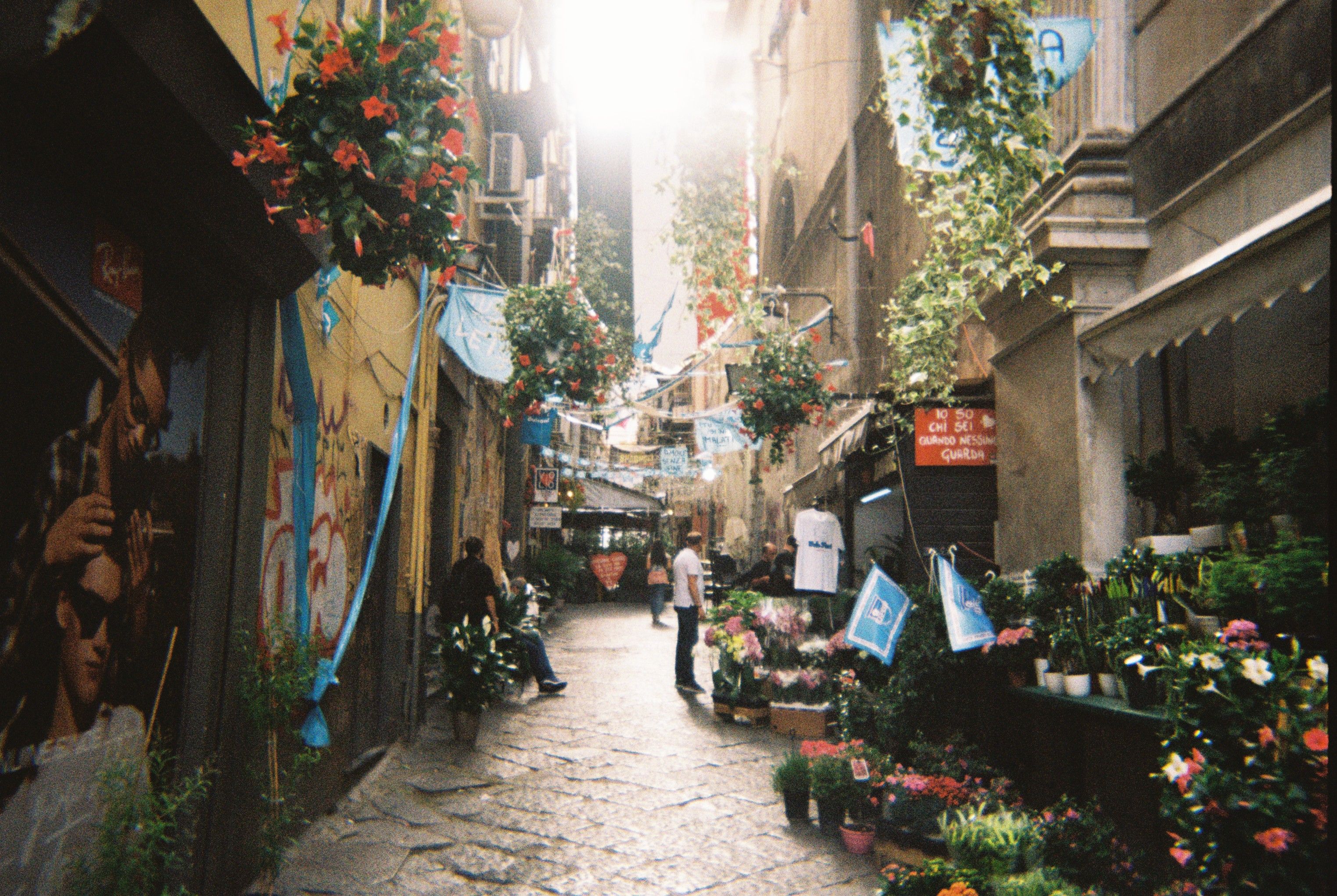 Vibrant alleyway with flowers, graffiti, and pedestrians under sunlight.