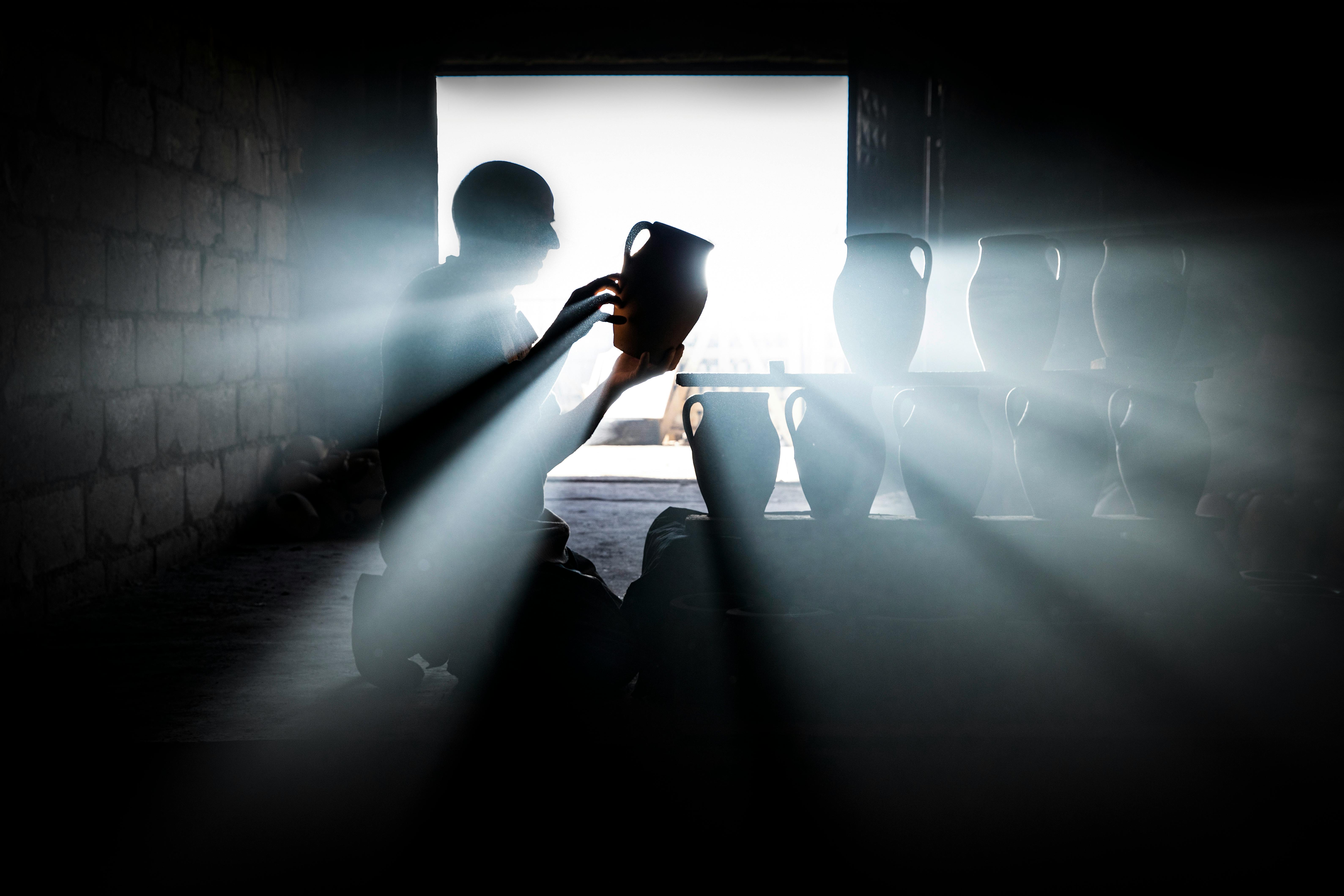 Gratis Dramatisk silhuett av en håndverker som vurderer keramikk med strøm av lysstråler i et rustikt verksted. Arkivbilde