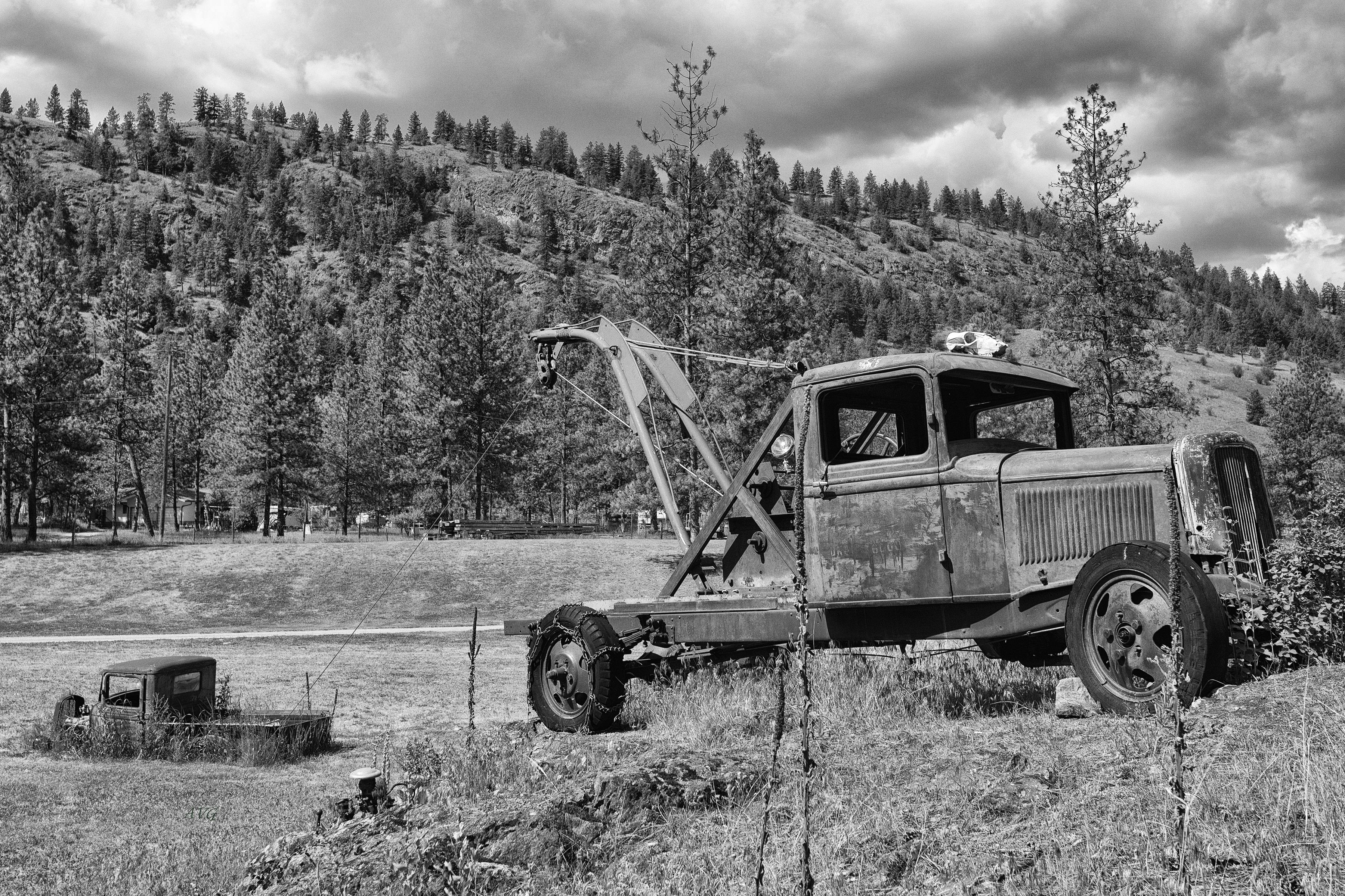 Black and white photo of a vintage tow truck in a rural landscape with mountains and pine trees.