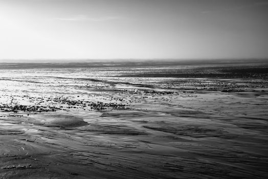 A dramatic black and white seascape photograph capturing a serene low tide with expansive mudflats.