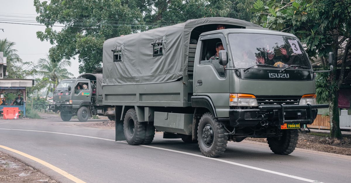 Military trucks traveling on a rural road surrounded by trees and local buildings.
