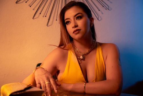 Free A woman sits indoors wearing a yellow dress and gold jewelry under warm lighting. Stock Photo