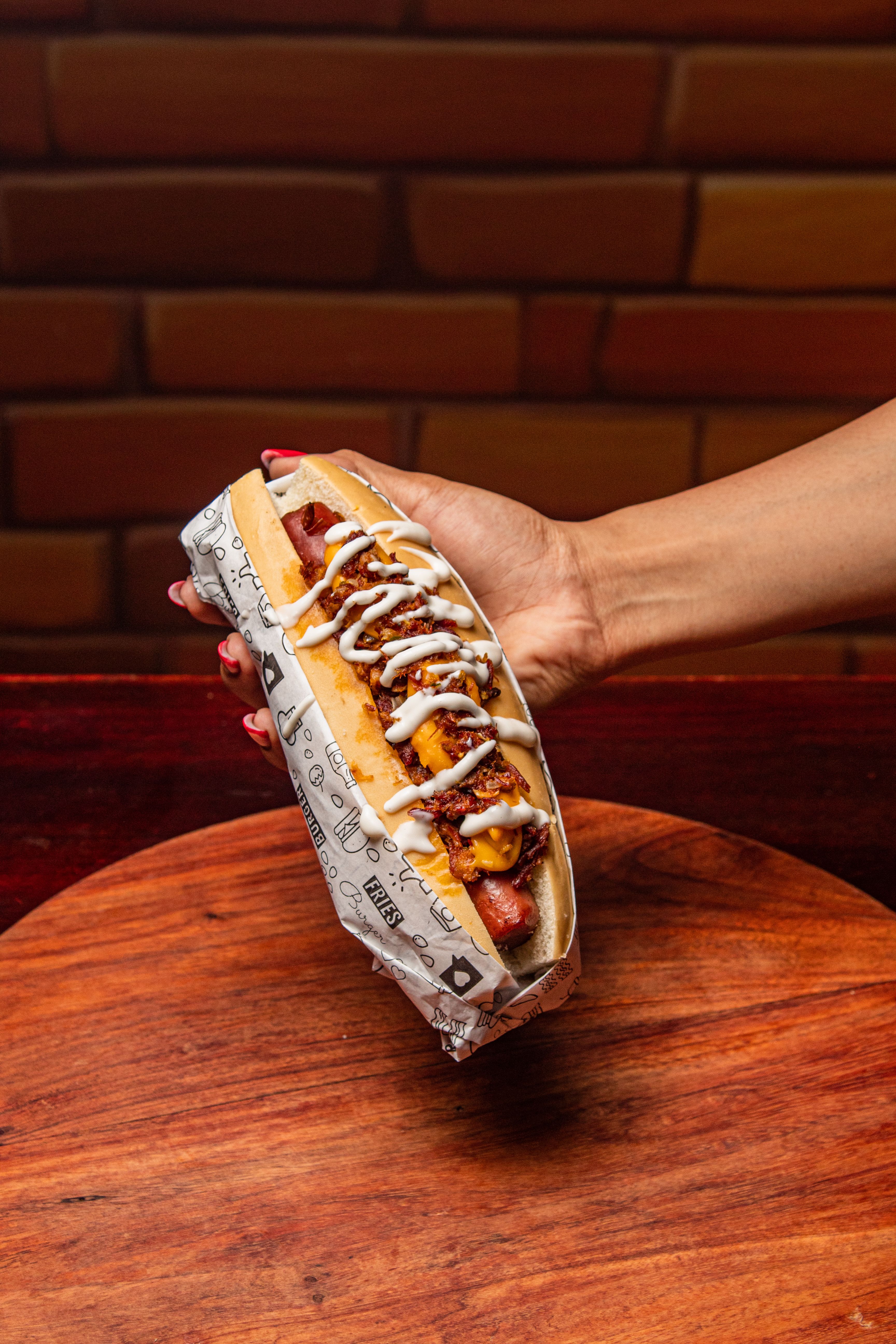 Appetizing hot dog with toppings and sauces held by a hand against a warm background.