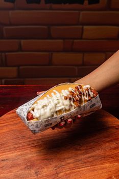 A hand presents a loaded hot dog with toppings and sauce, wrapped in paper, on a wooden board.