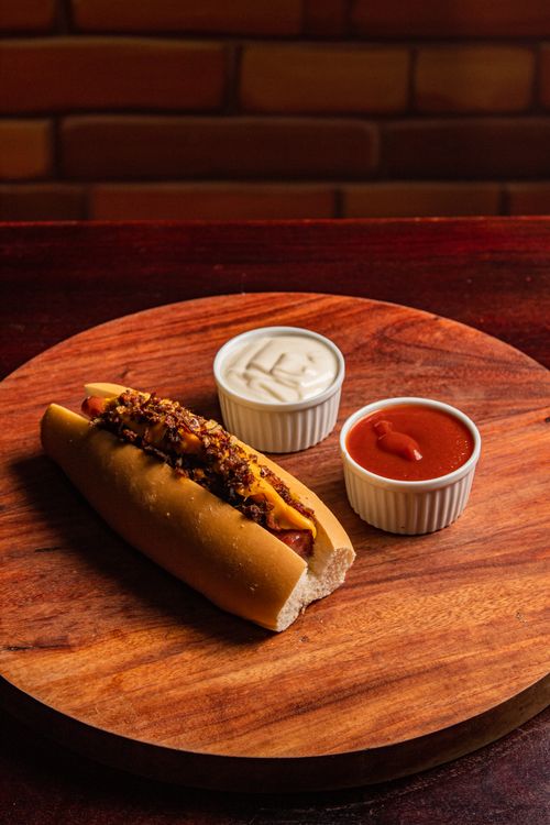 Free A delicious gourmet hot dog with chili, cheese, and sauces, styled on a wooden platter. Stock Photo