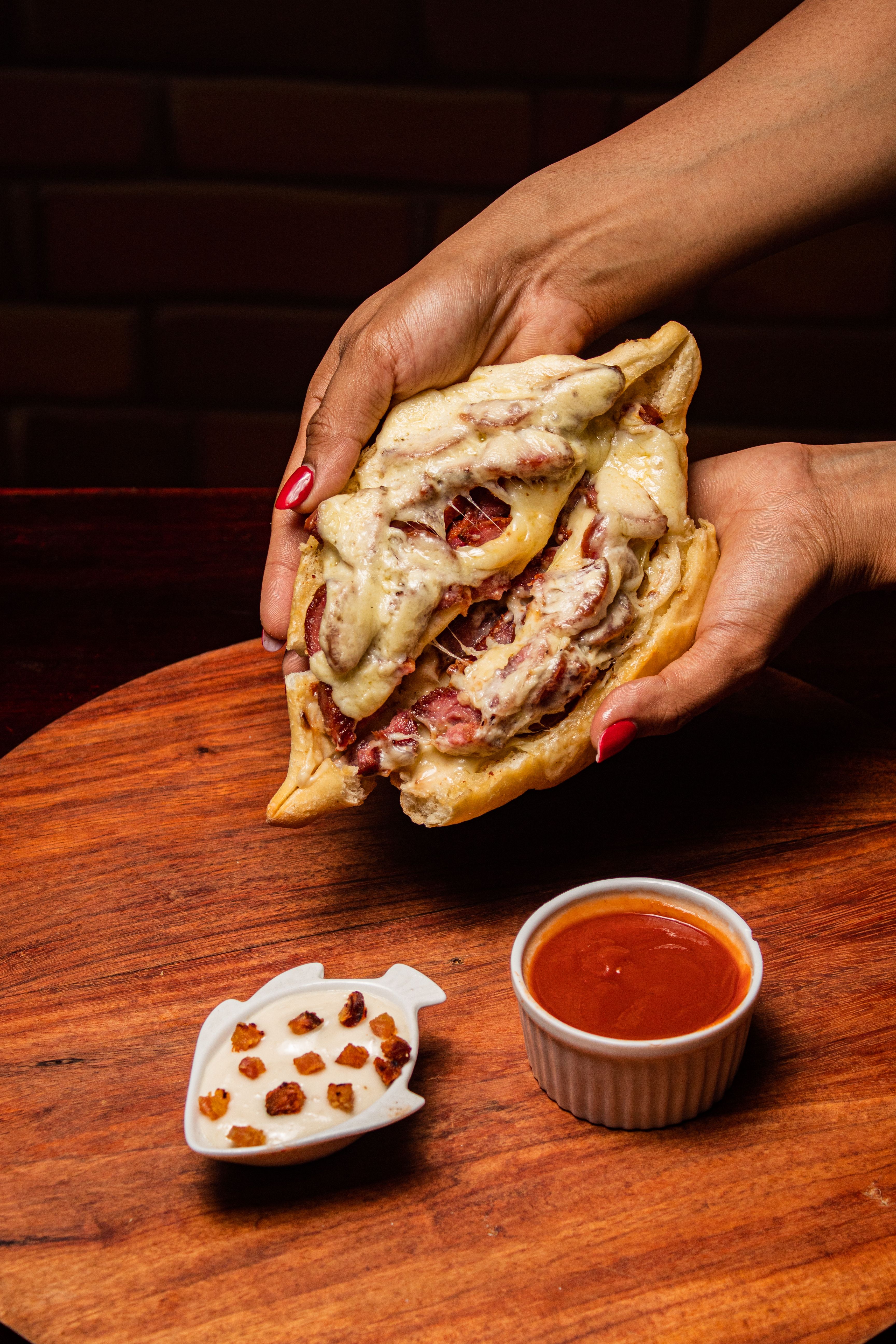 A close-up of hands holding a cheesy sandwich with sauces on a wooden board. Perfect for food lovers.