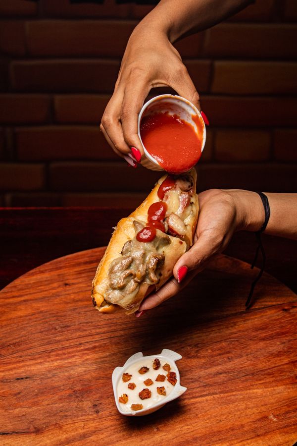 Imagem em close-up de um cachorro-quente sendo coberto com molho de tomate manualmente sobre uma mesa de madeira.