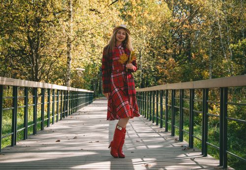 Free A woman in a red plaid dress holds yellow leaves while walking on a wooden bridge in a vibrant autumn setting. Stock Photo