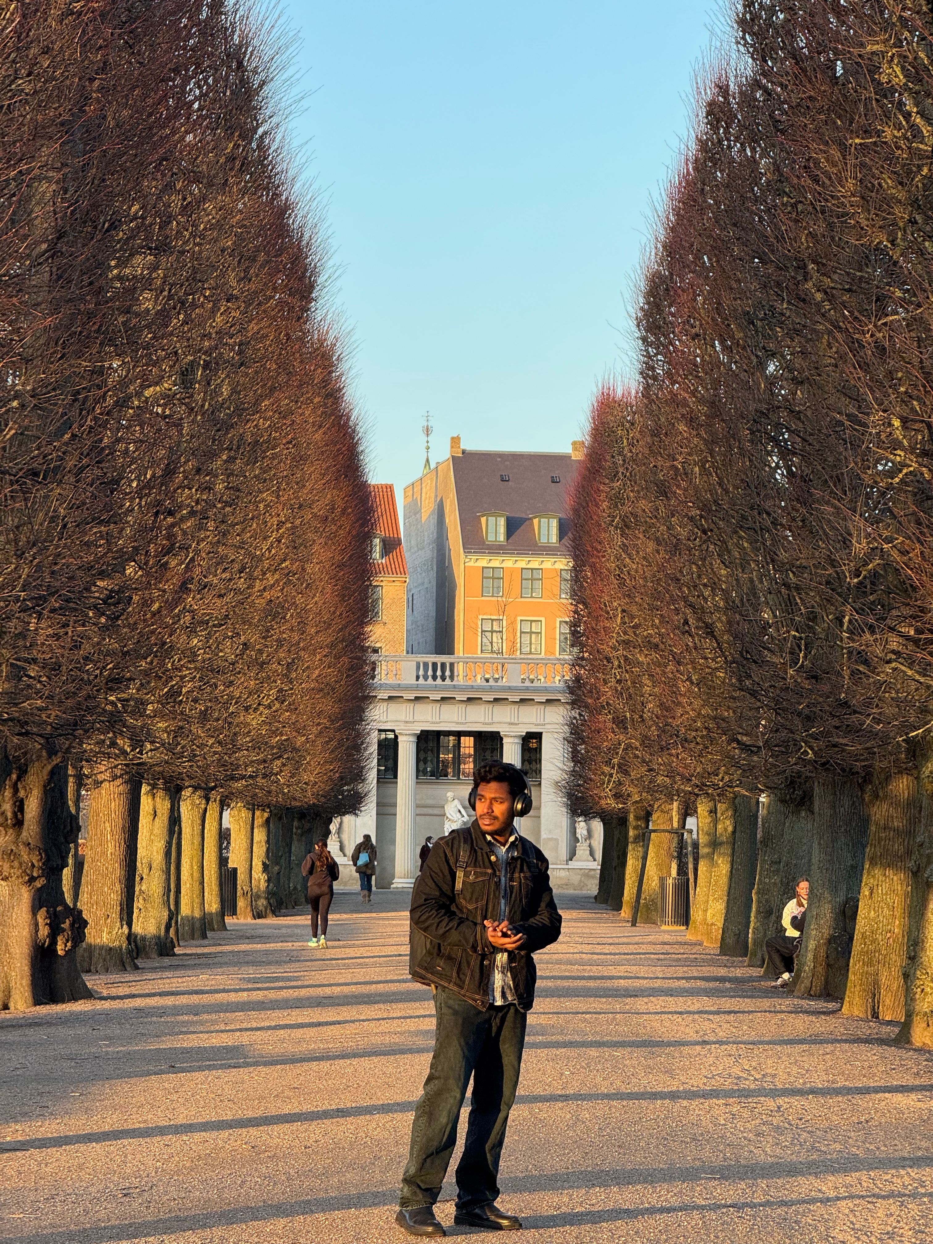 A young man walks through a tree-lined path in Copenhagen, surrounded by classic architecture.