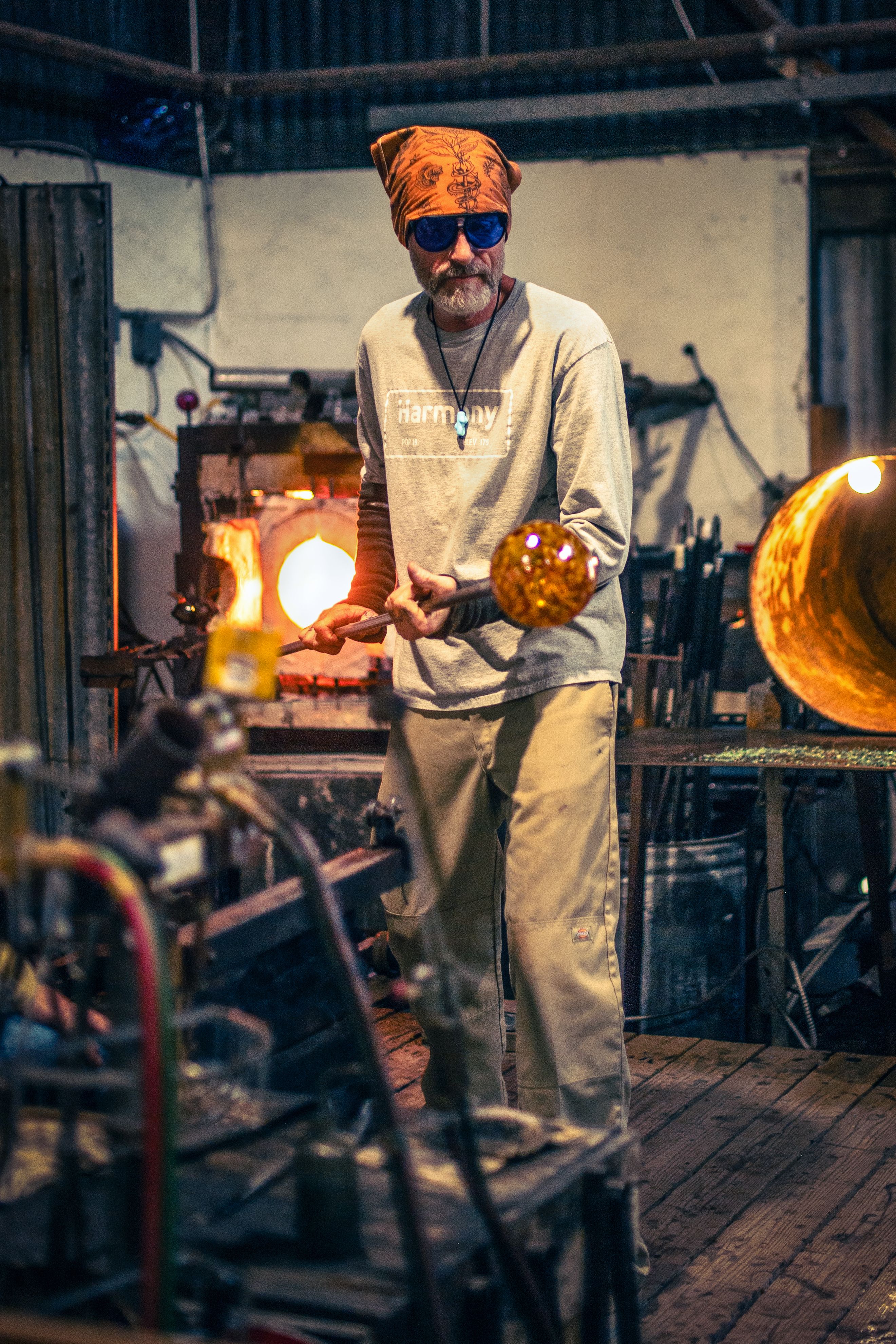 An artisan skillfully shapes molten glass in a Harmony, California workshop, surrounded by tools and a warm glow.