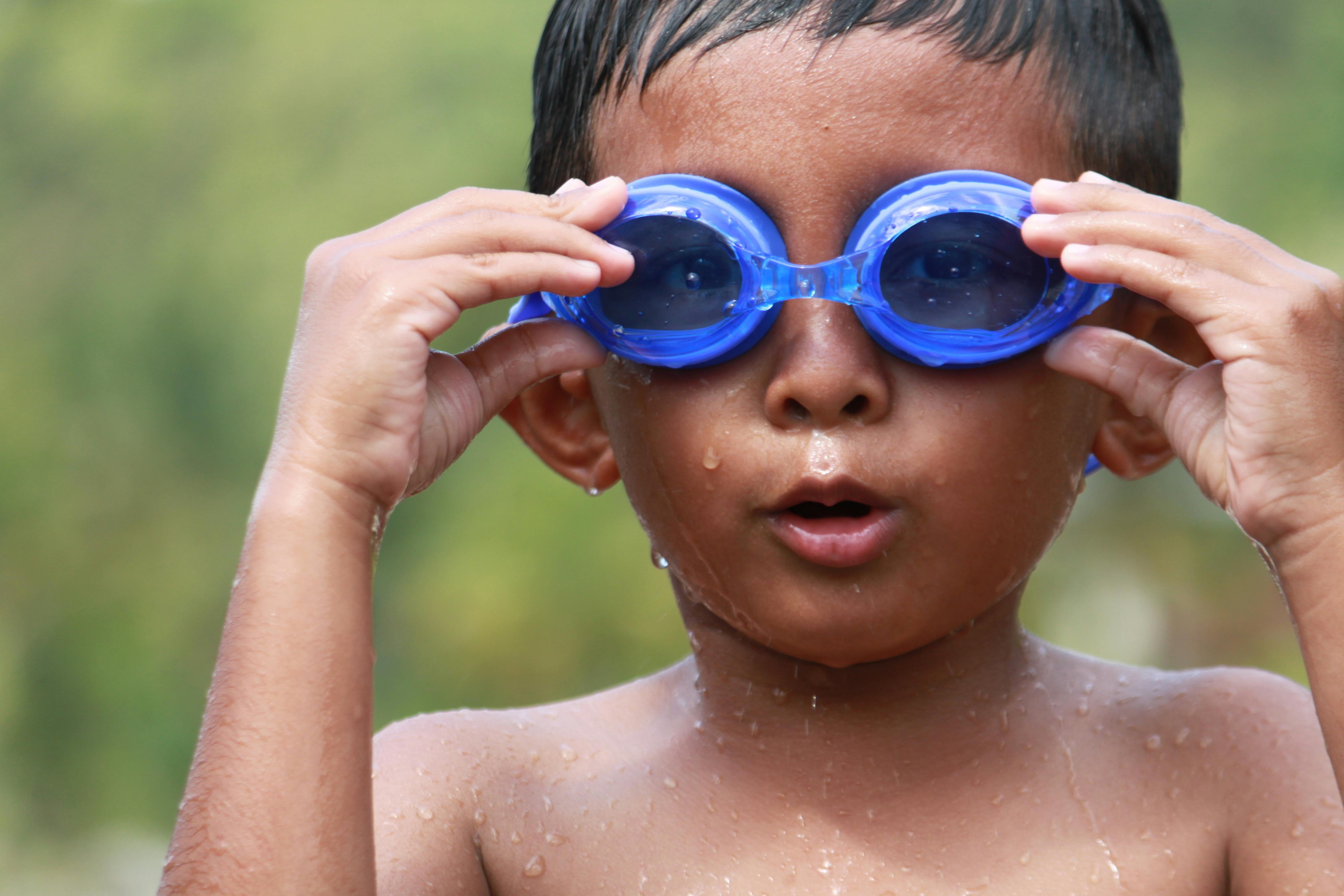 Ücretsiz Açık havada duran, mavi yüzme gözlüğü takmış küçük bir çocuk, teninde su damlacıklarıyla şaşkın bir ifadeyle bakıyor. Stok Fotoğraflar