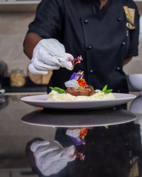 Chef carefully garnishing a gourmet beef steak with edible flowers and greens on a plate of mashed potatoes.