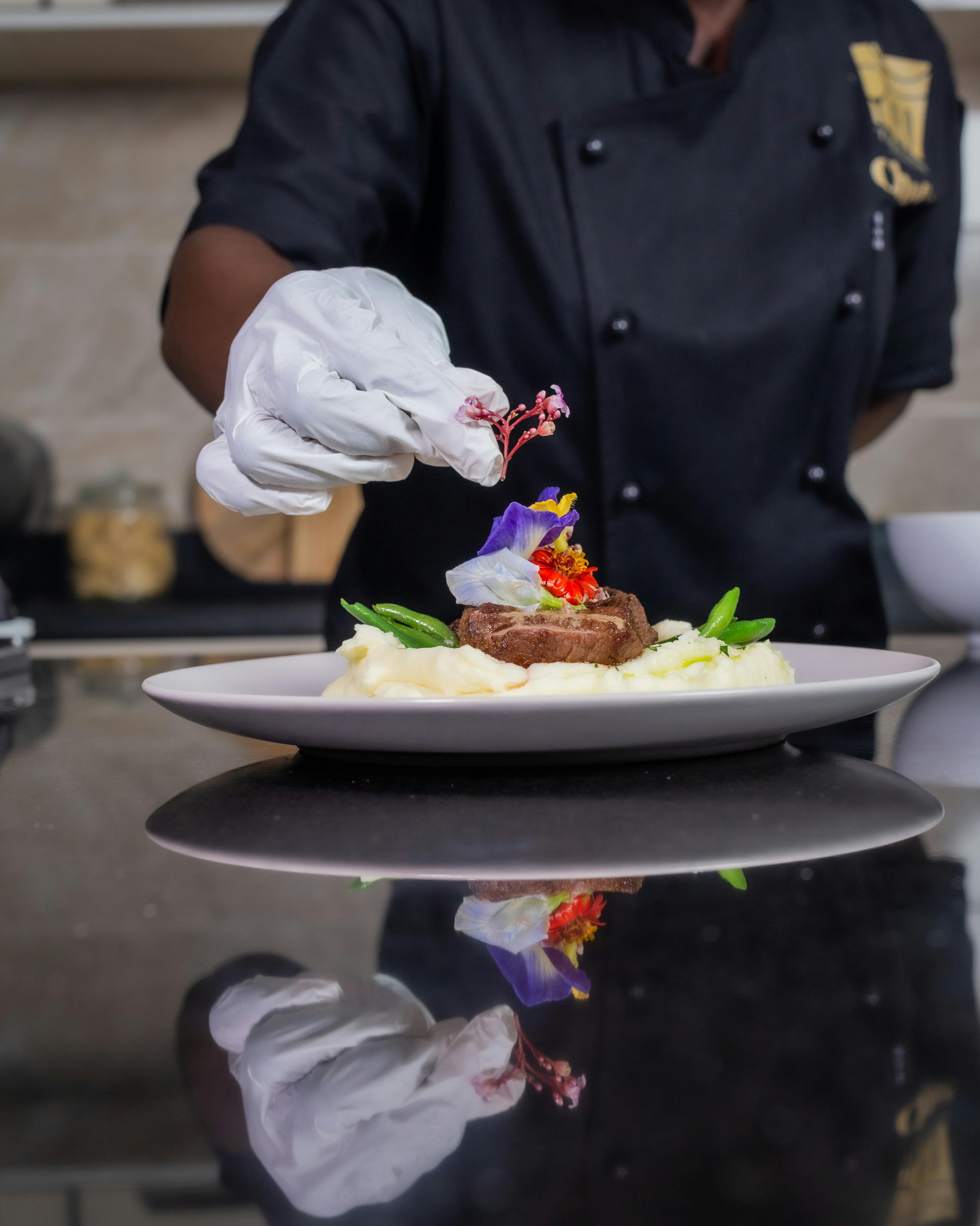 Gratis Kok pynter omhyggeligt en gourmet oksebøf med spiselige blomster og grøntsager på en tallerken med kartoffelmos. Lagerfoto