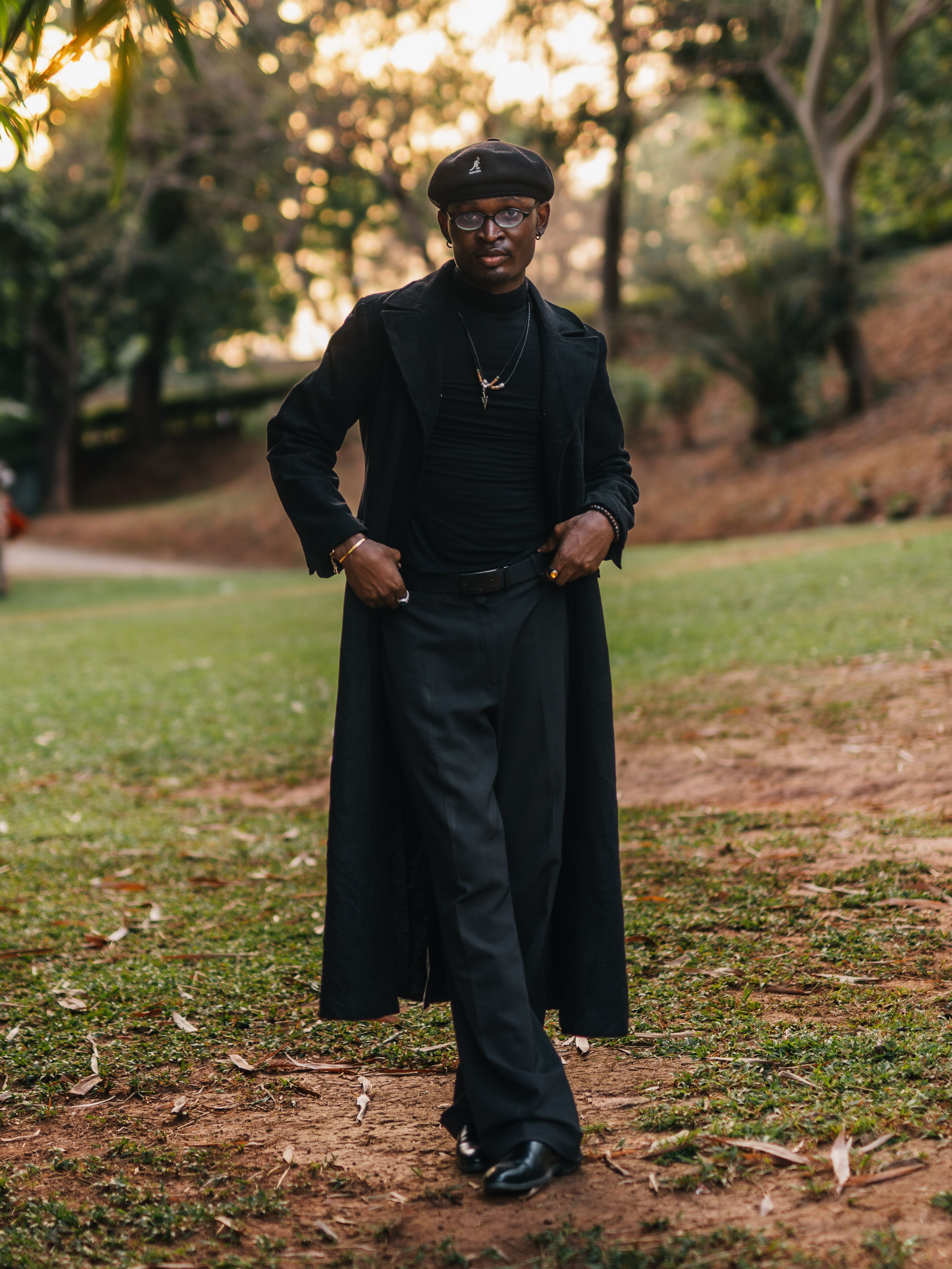 A fashionable man in a city park in Abuja, wearing a black coat at sunset.