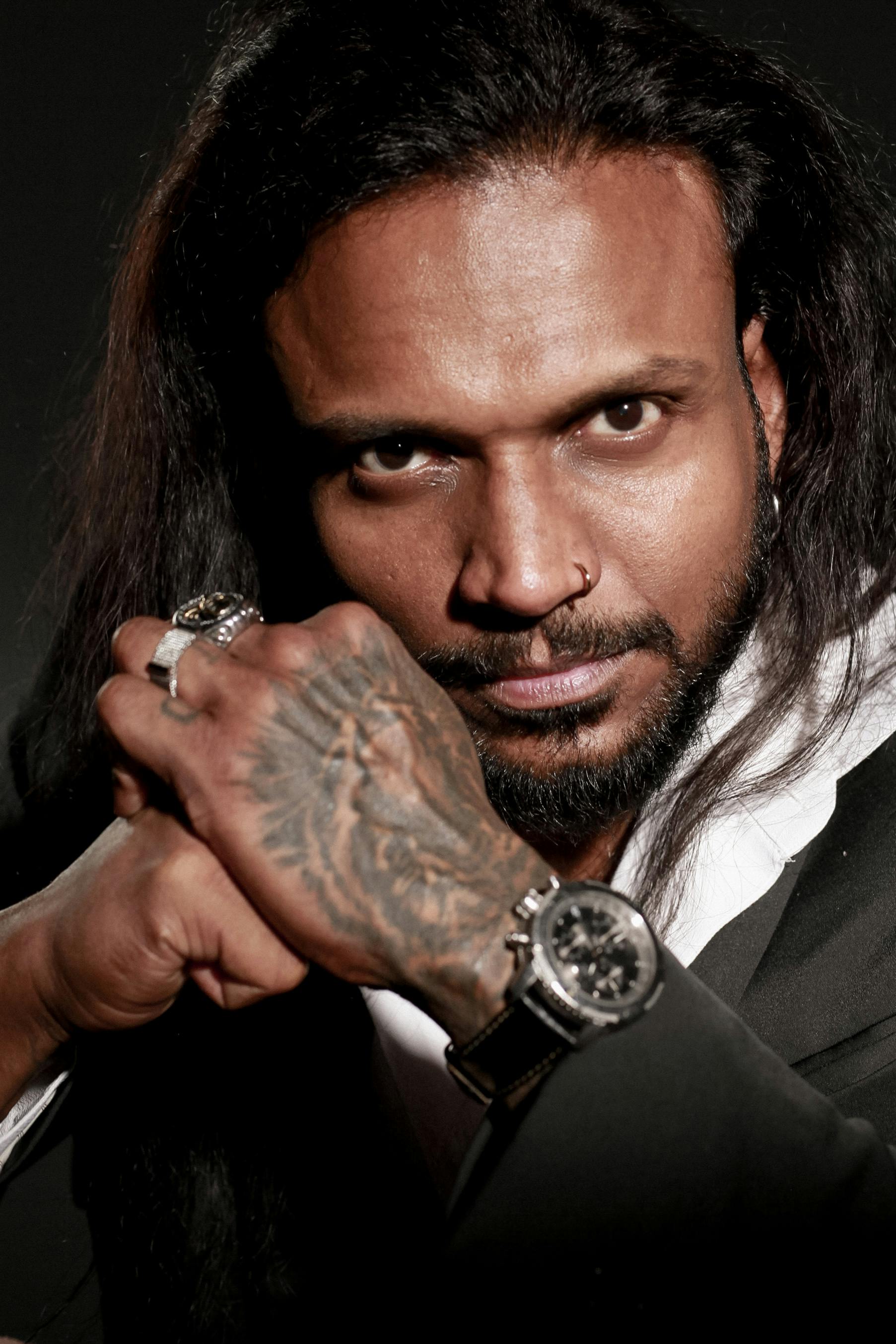 Close-up portrait of a man with tattoos, rings, and a watch. Intense gaze and moody lighting create a compelling image.