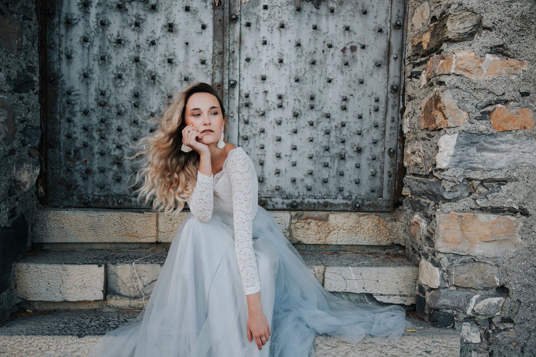 Woman in elegant lace dress sitting pensively by rustic iron door.
