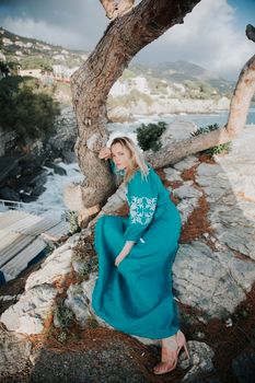 Elegant woman in blue dress poses by a tree on a rocky coastal landscape.