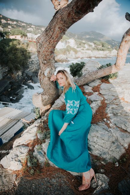Elegant woman in blue dress poses by a tree on a rocky coastal landscape.