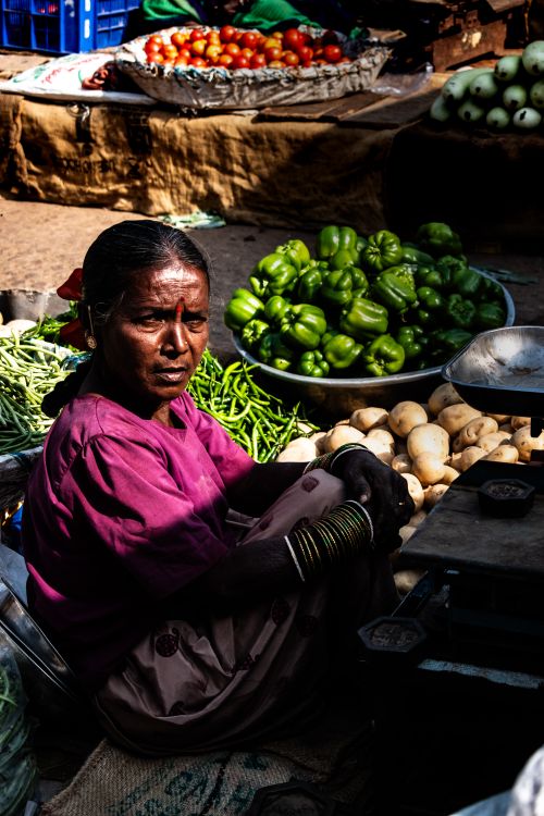 Free A woman vendor at an outdoor market sitting among fresh vegetables like bell peppers and potatoes. Stock Photo