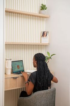 A young woman working on a laptop in a modern, stylish home office setting with shelves and plants.