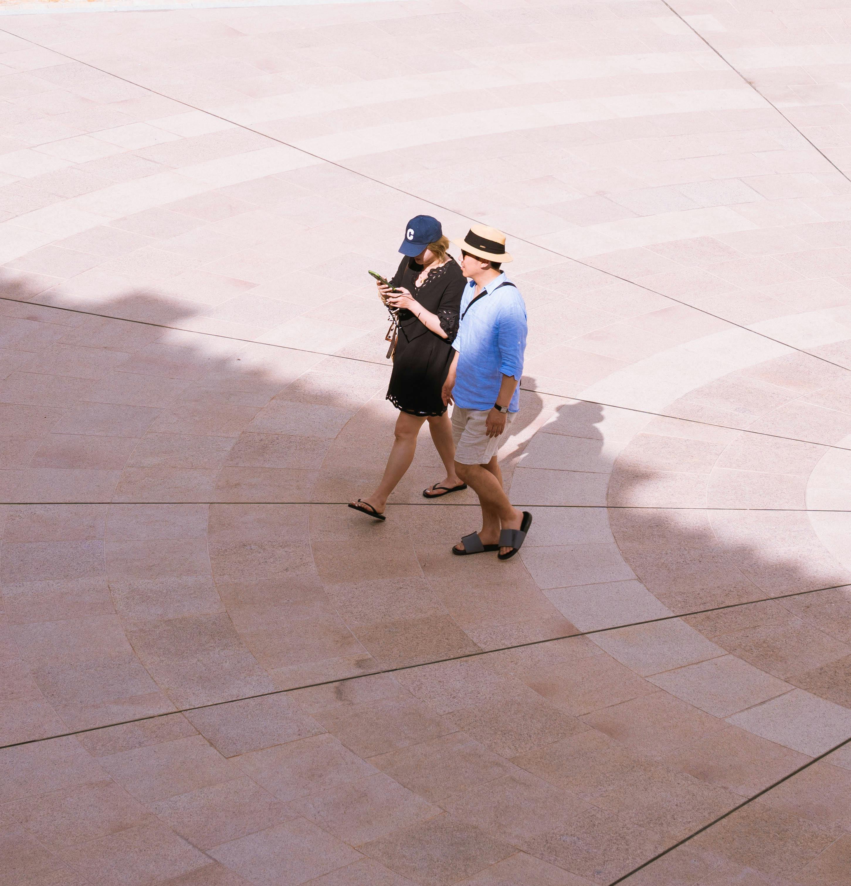 Gratuit Un couple se promène dans une cour pavée de pierres à motifs, sous un soleil éclatant qui projette des ombres. Photos