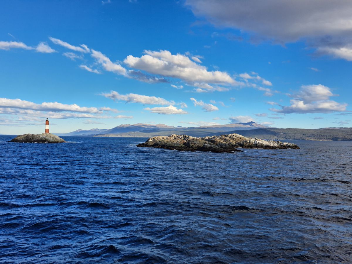 A lighthouse on a low rocky islet in a wide blue channel under scattered cloud.