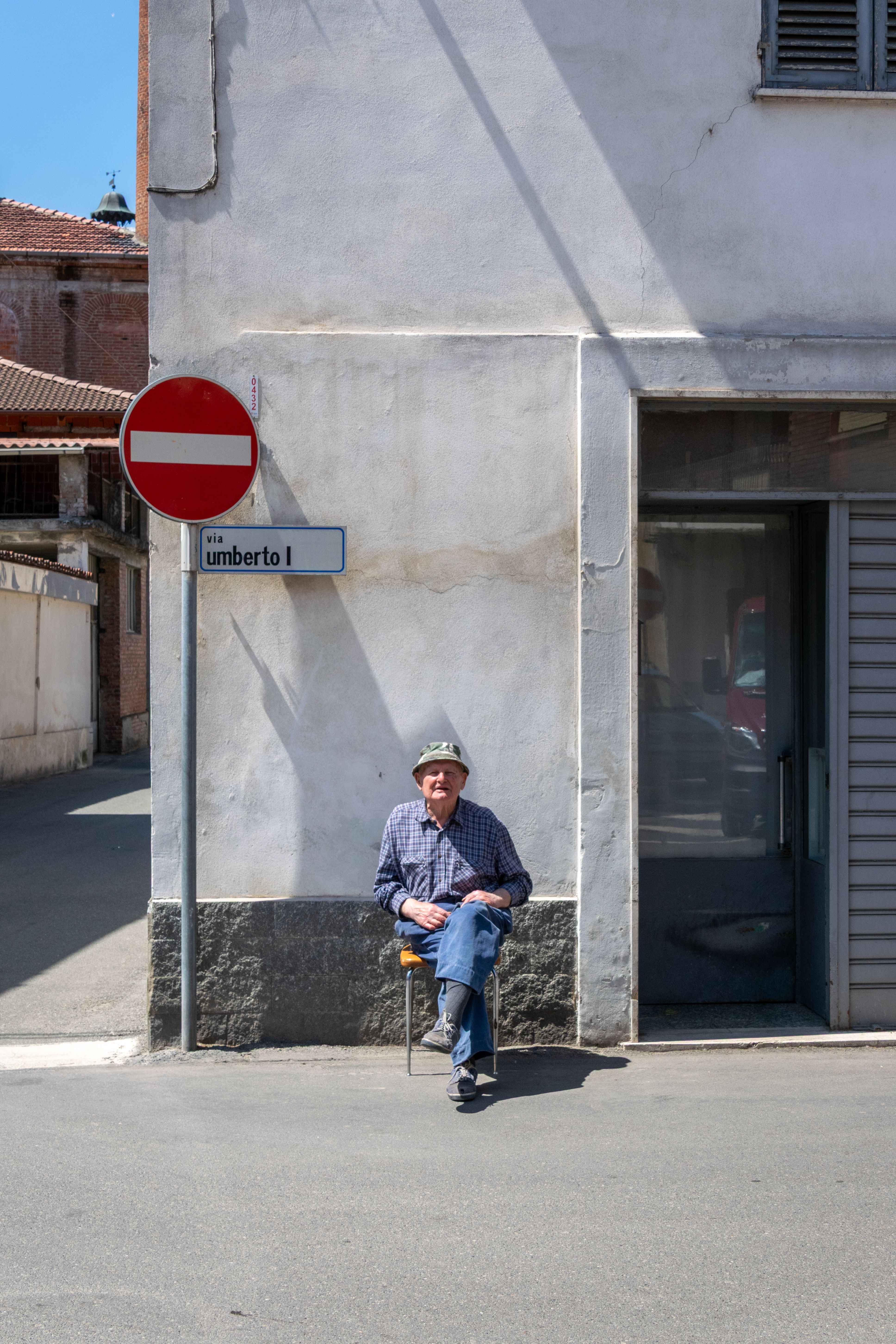 Elderly man sitting by a street sign, relaxes in the sun in Castellazzo Bormida, Italy.