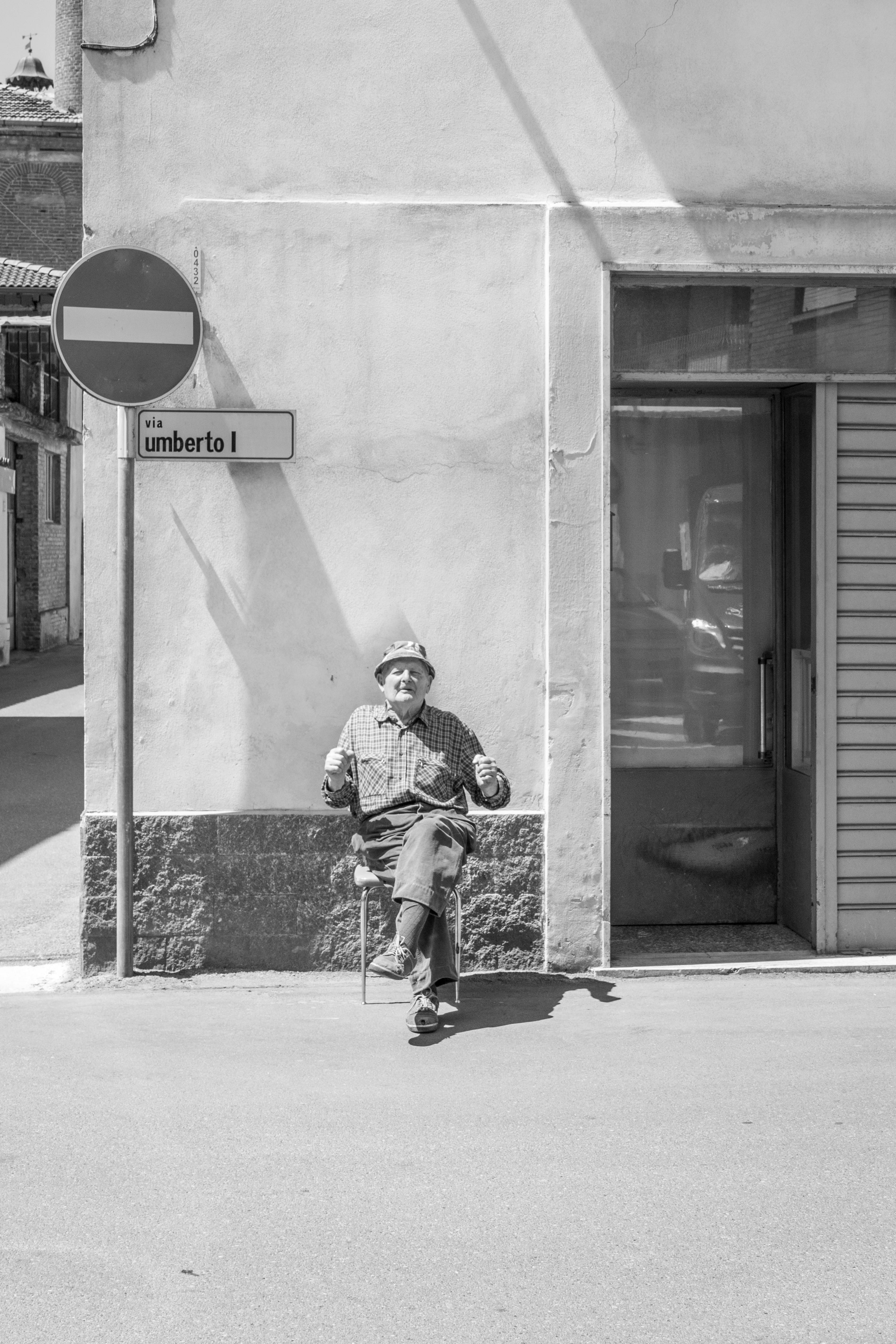 bezplatná Černobílá fotografie staršího muže sedícího venku na ulici Via Umberto I. v Castellazzo Bormida v Itálii. Základová fotografie