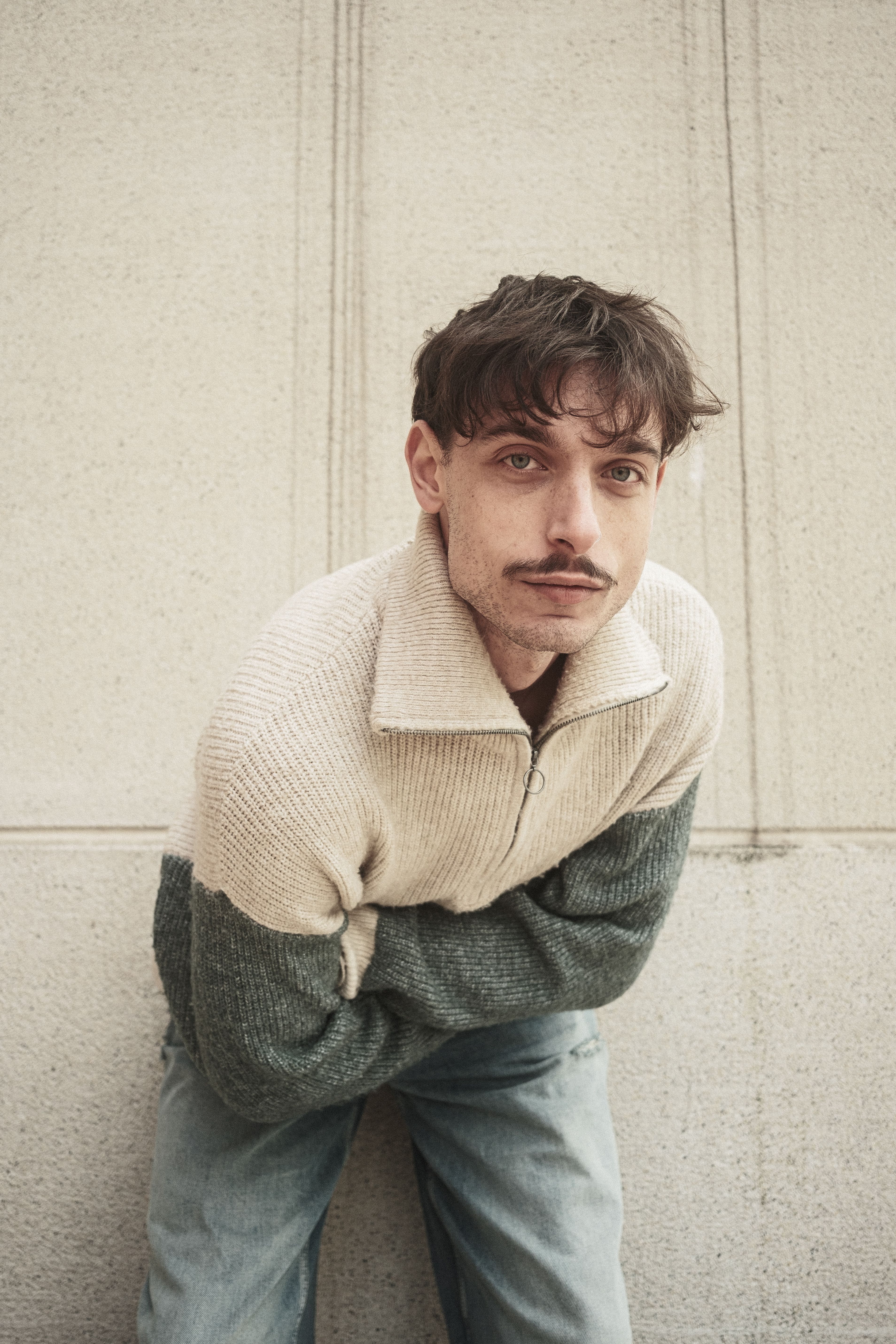 Stylish young man with mustache poses against a concrete wall.