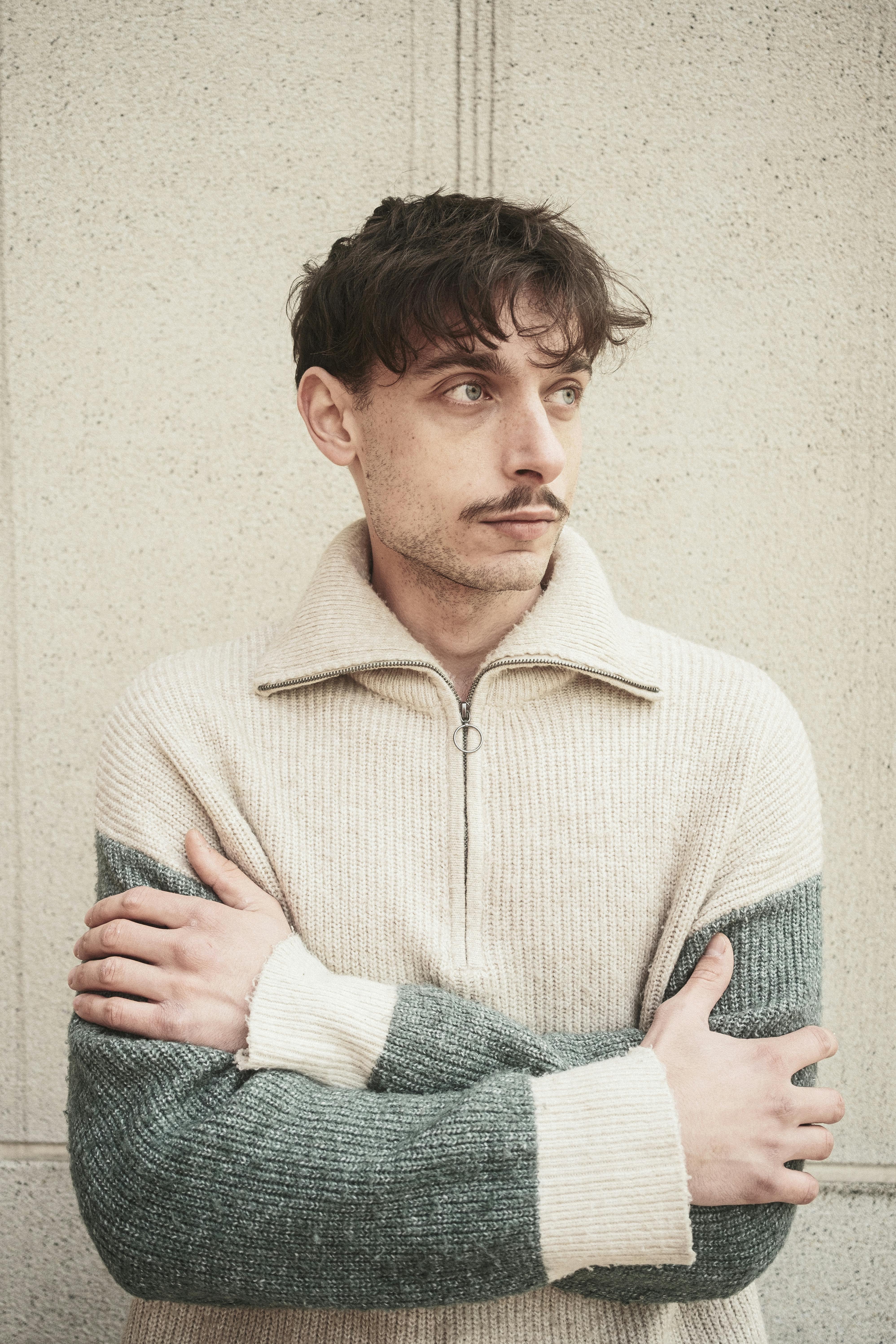Free A man in a cozy sweater stands against a textured wall, looking contemplative. Stock Photo