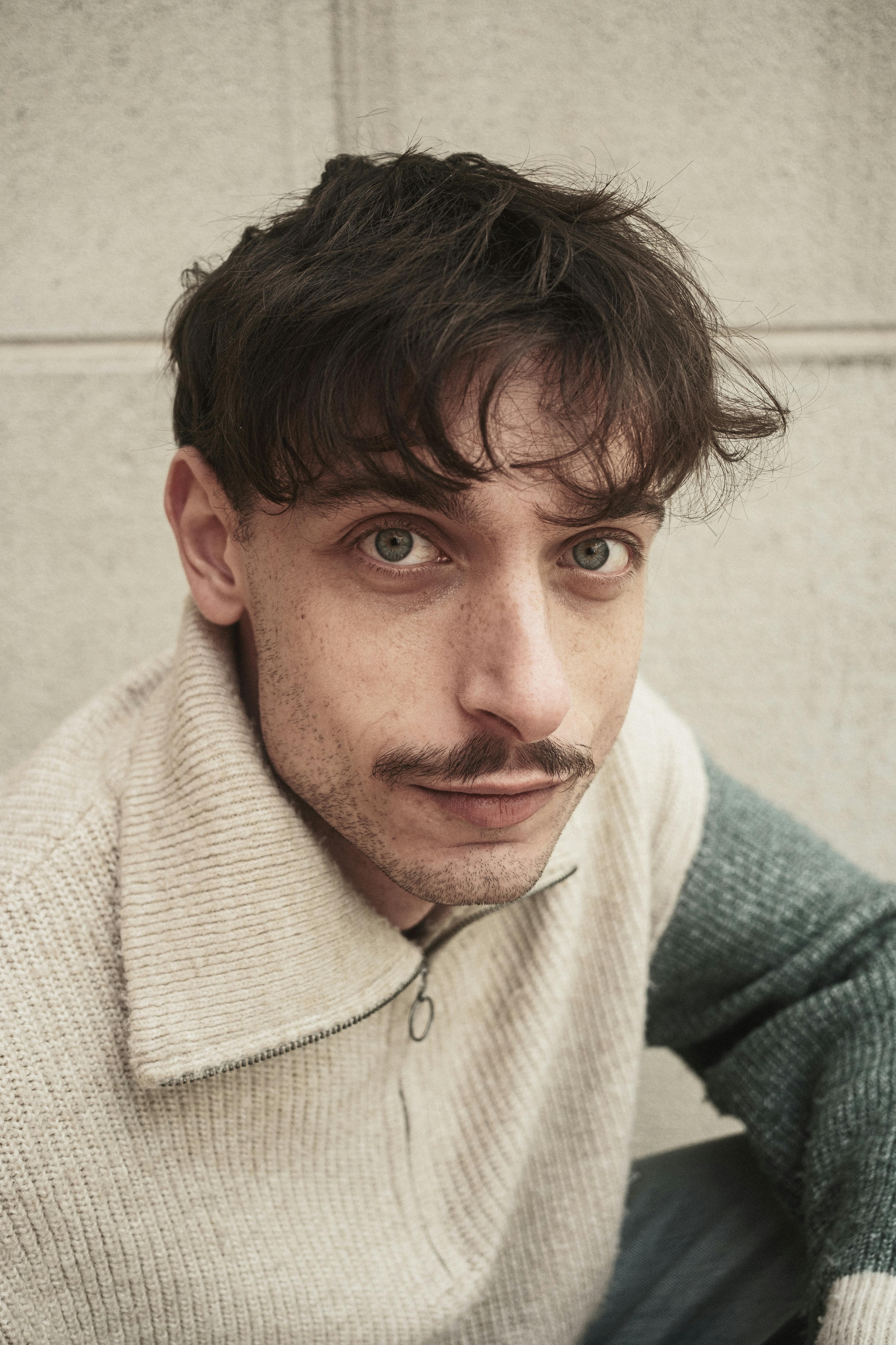 Close-up portrait of a young adult male with a mustache, wearing a sweater.