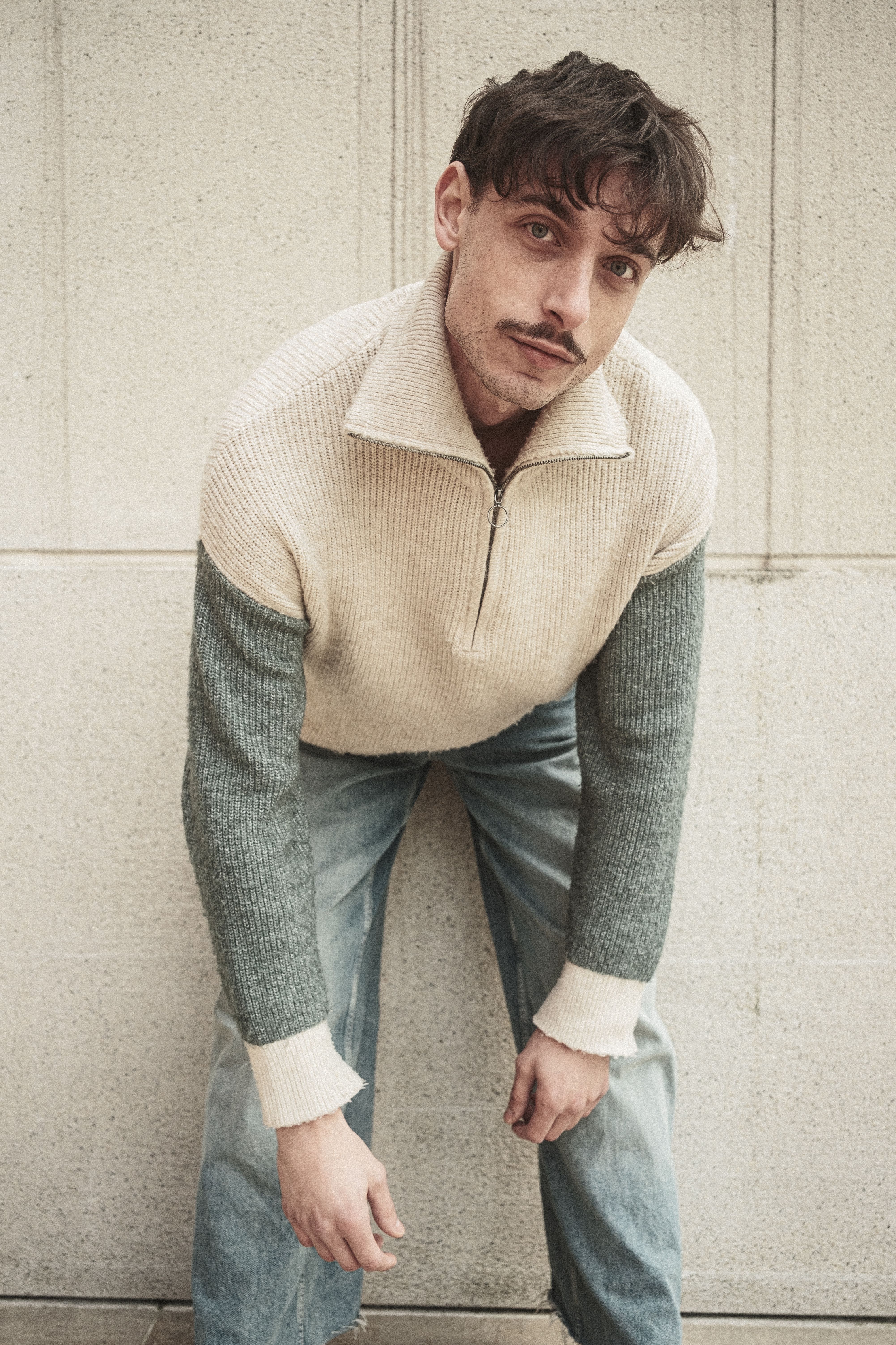 Fashion-forward young man in casual outfit leaning against a textured wall.