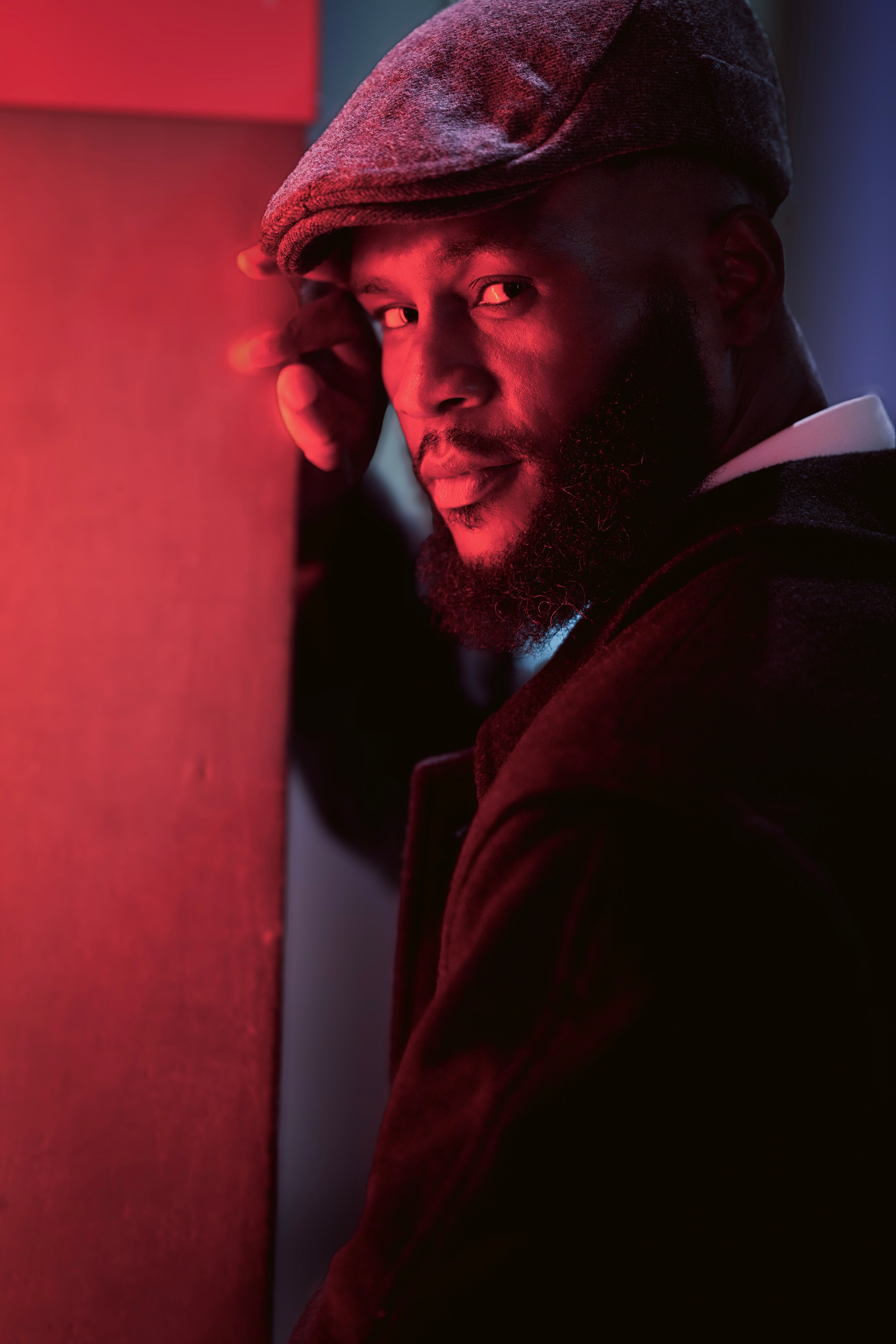 Gratis Un ritratto drammatico di un uomo illuminato di rosso, che indossa un berretto piatto e un cappotto, e che emana un'atmosfera malinconica. Foto a disposizione