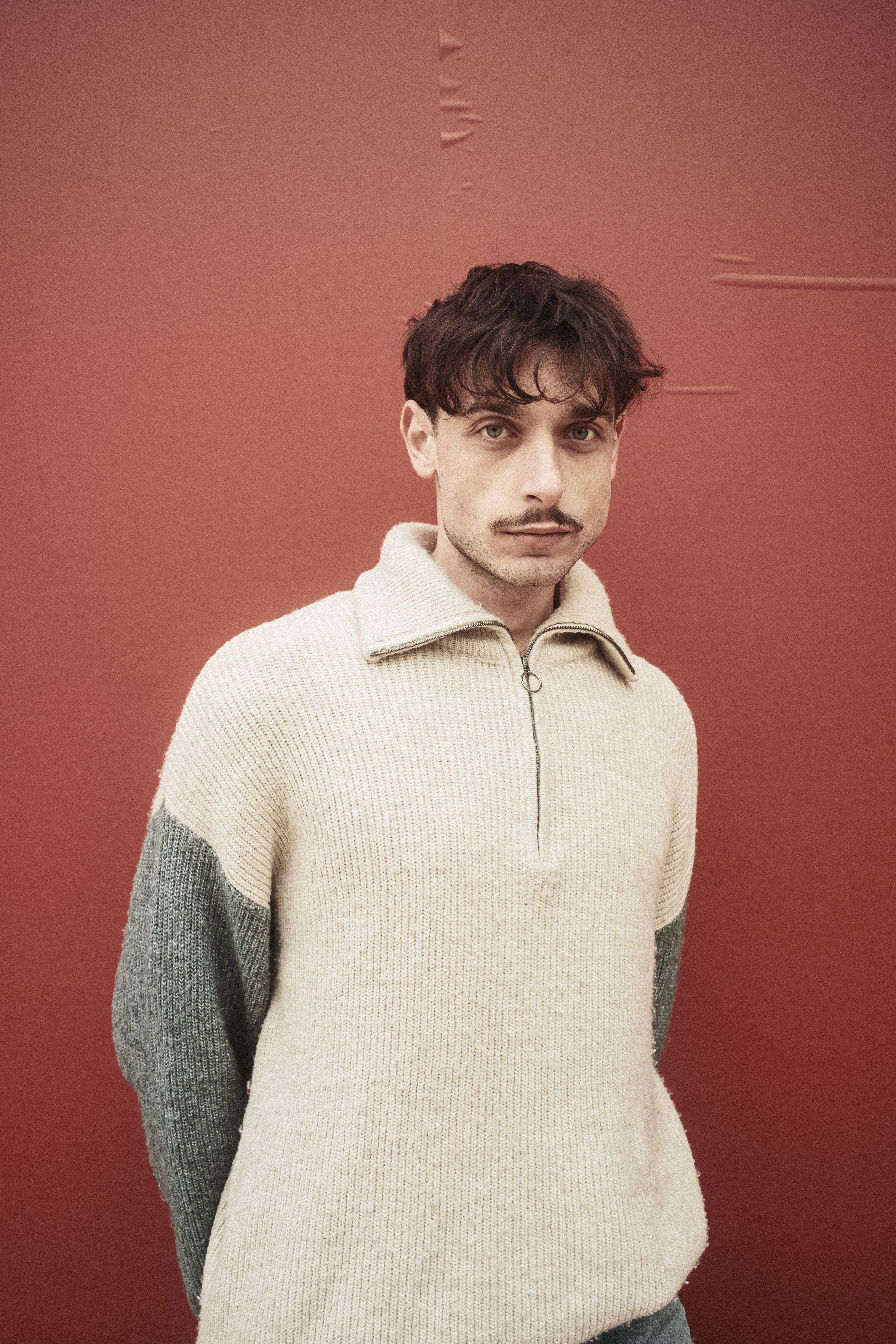 Stylish man in a cozy sweater posing in front of a textured red wall indoors.