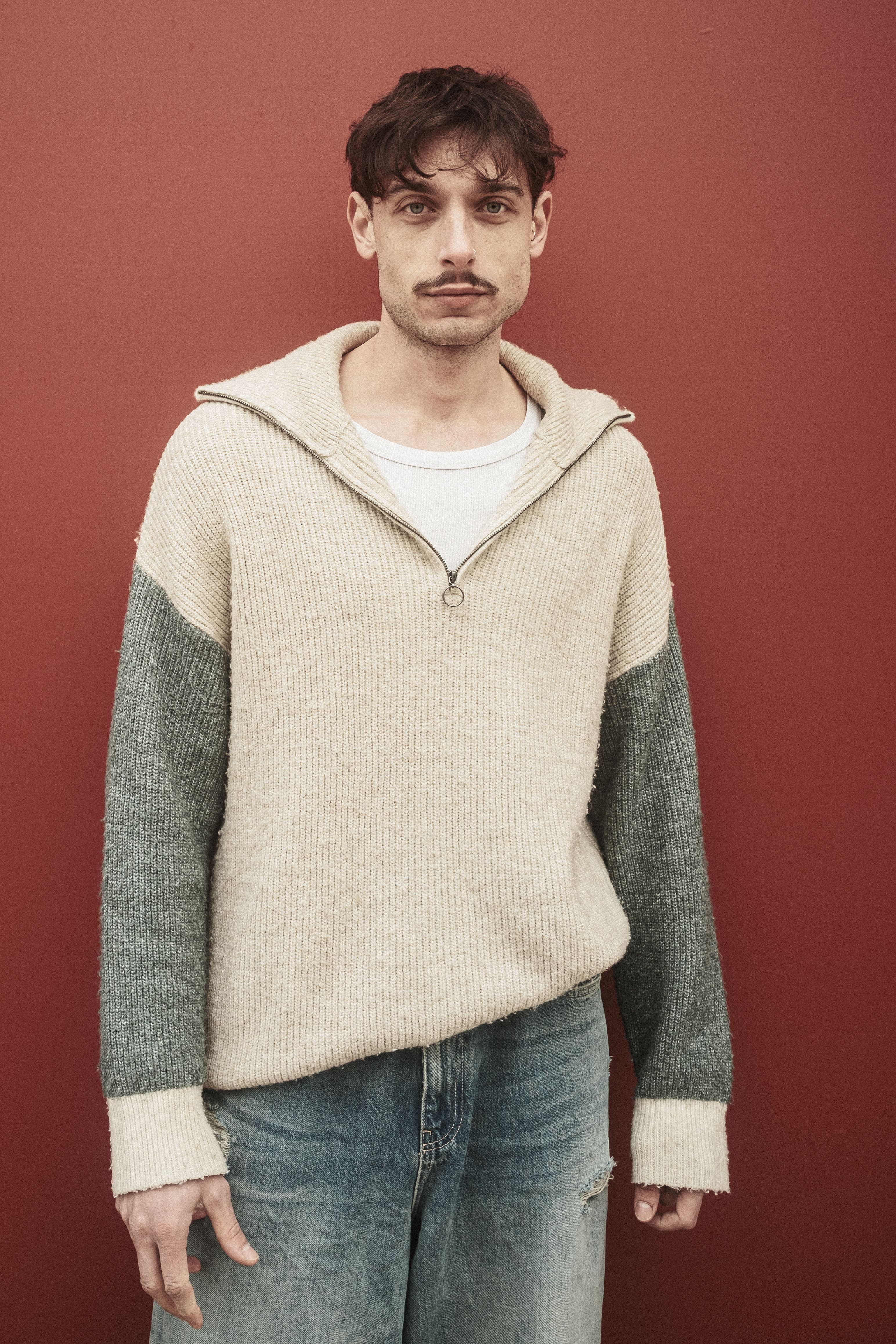 A young man in a casual sweater poses against a red backdrop.