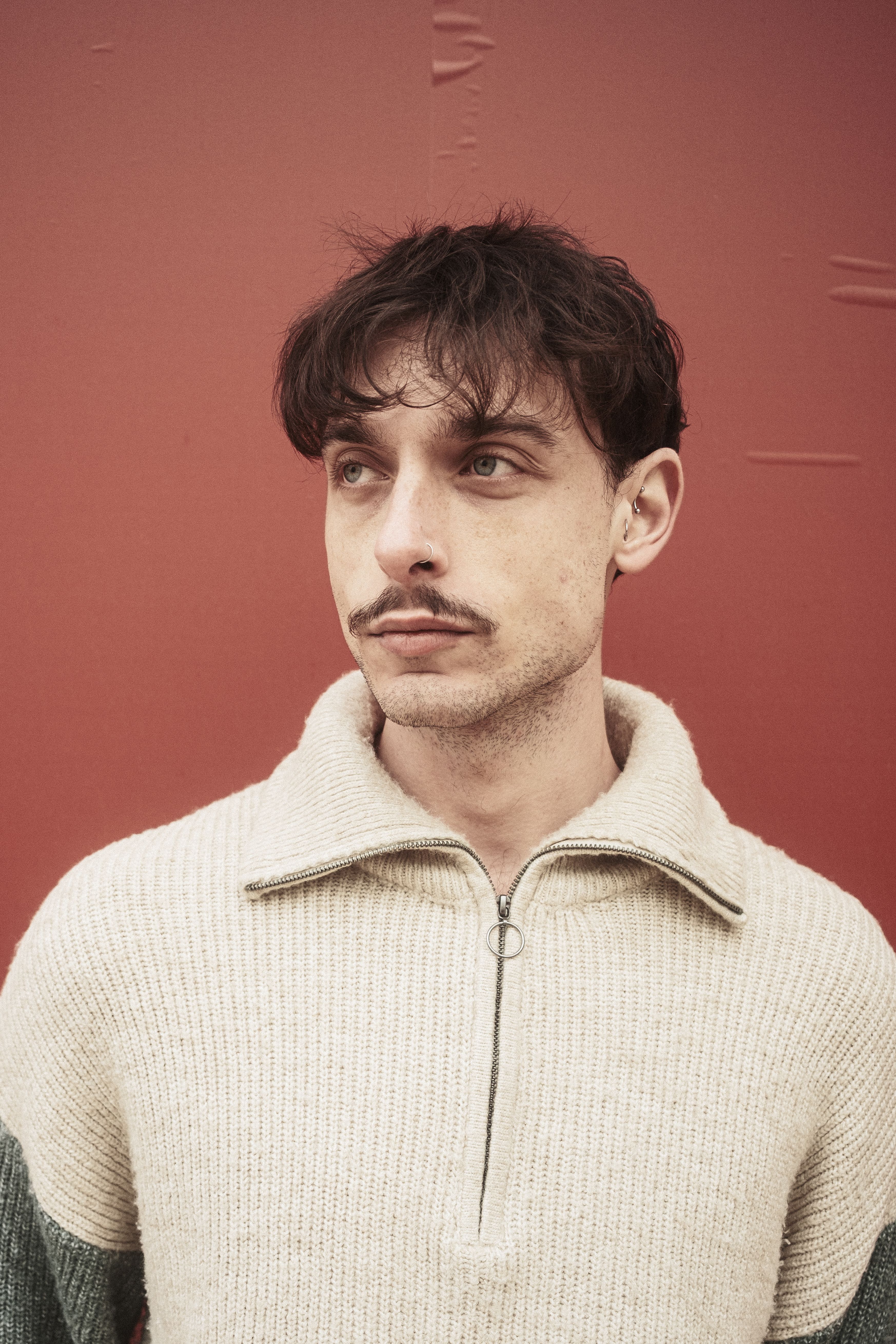 A man in a sweater stands pensively against a red background, capturing a moment of introspection.