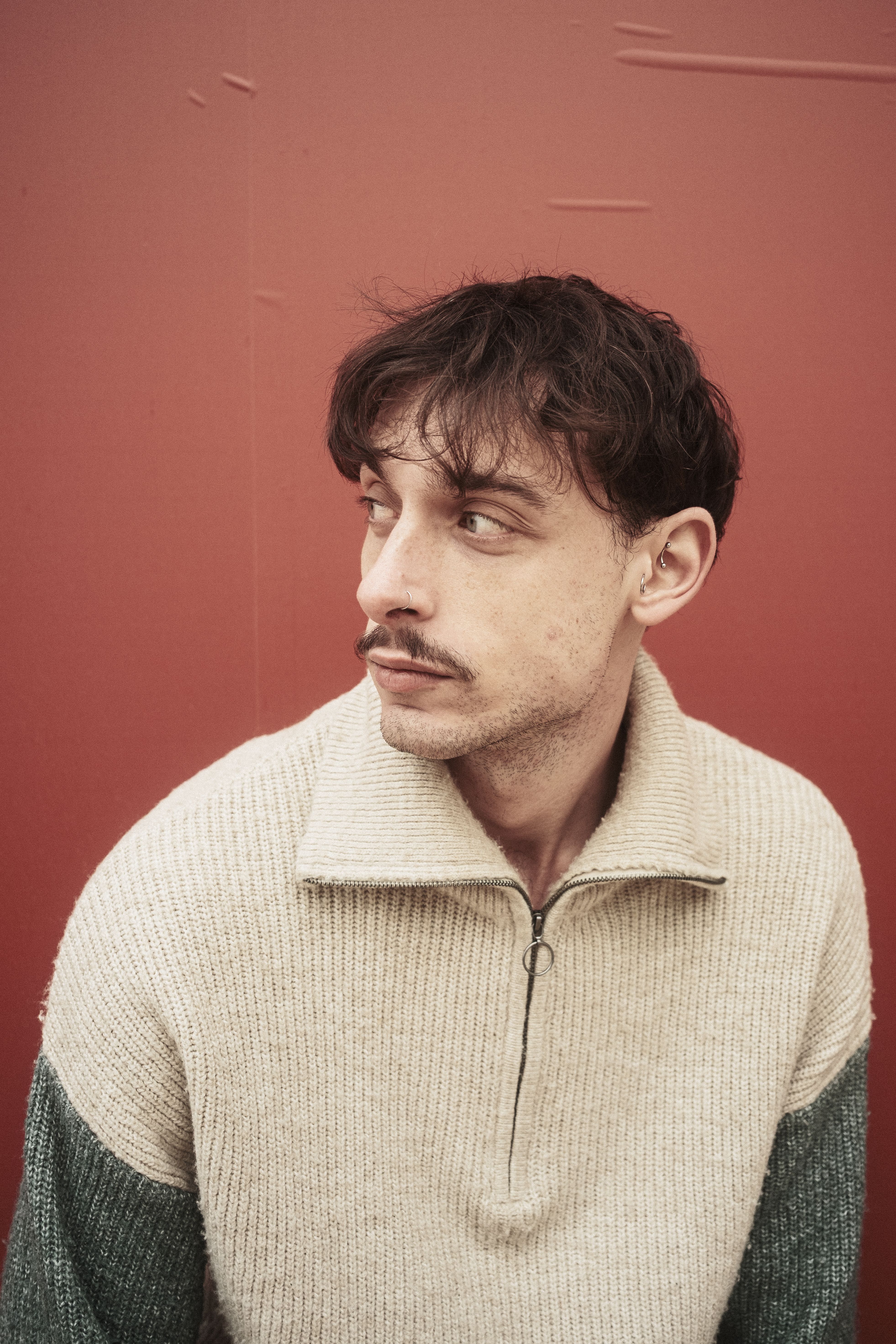 Thoughtful man in knit sweater posed against red backdrop, looking away.