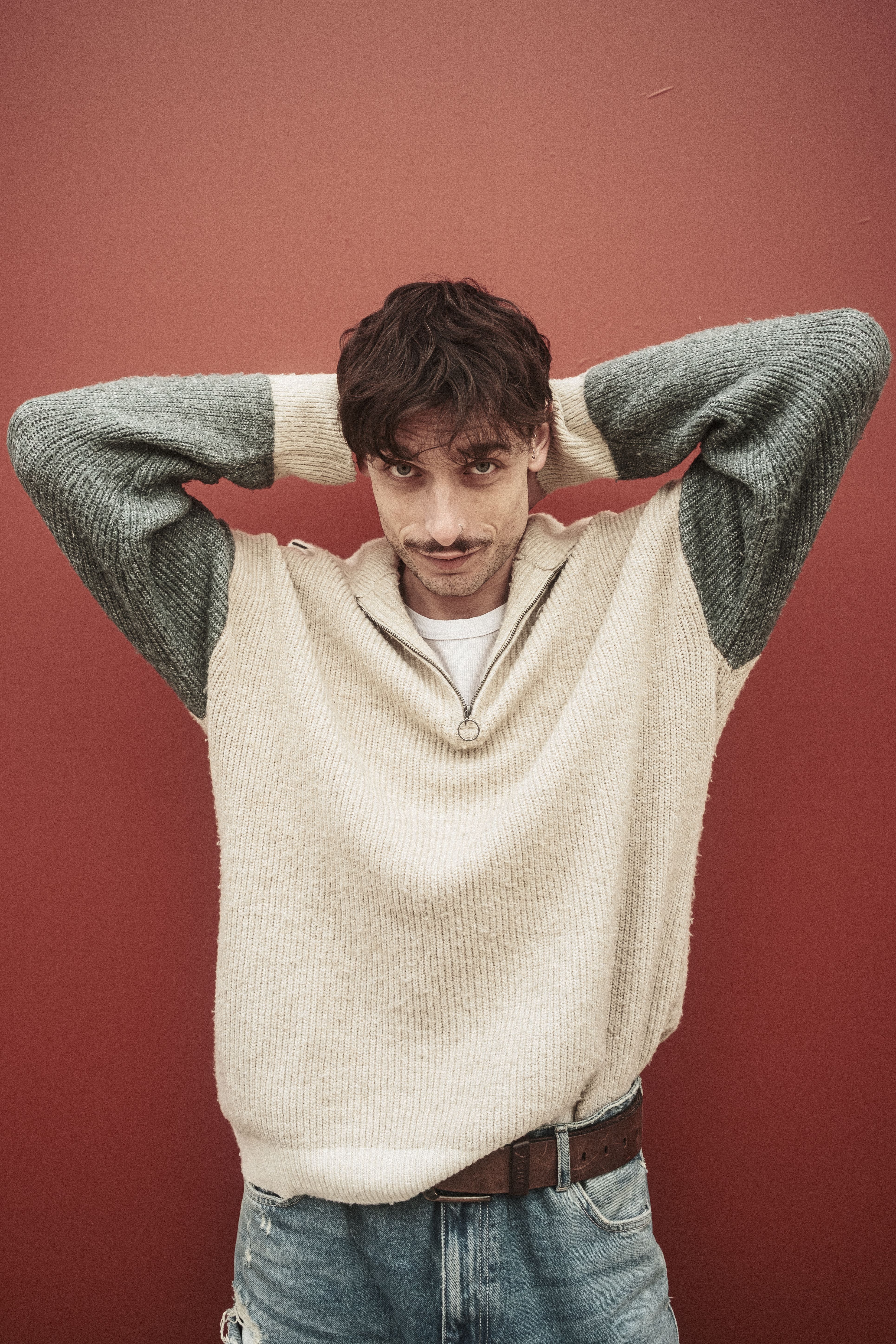 Man in a sweater and jeans posing with hands behind head against red wall.