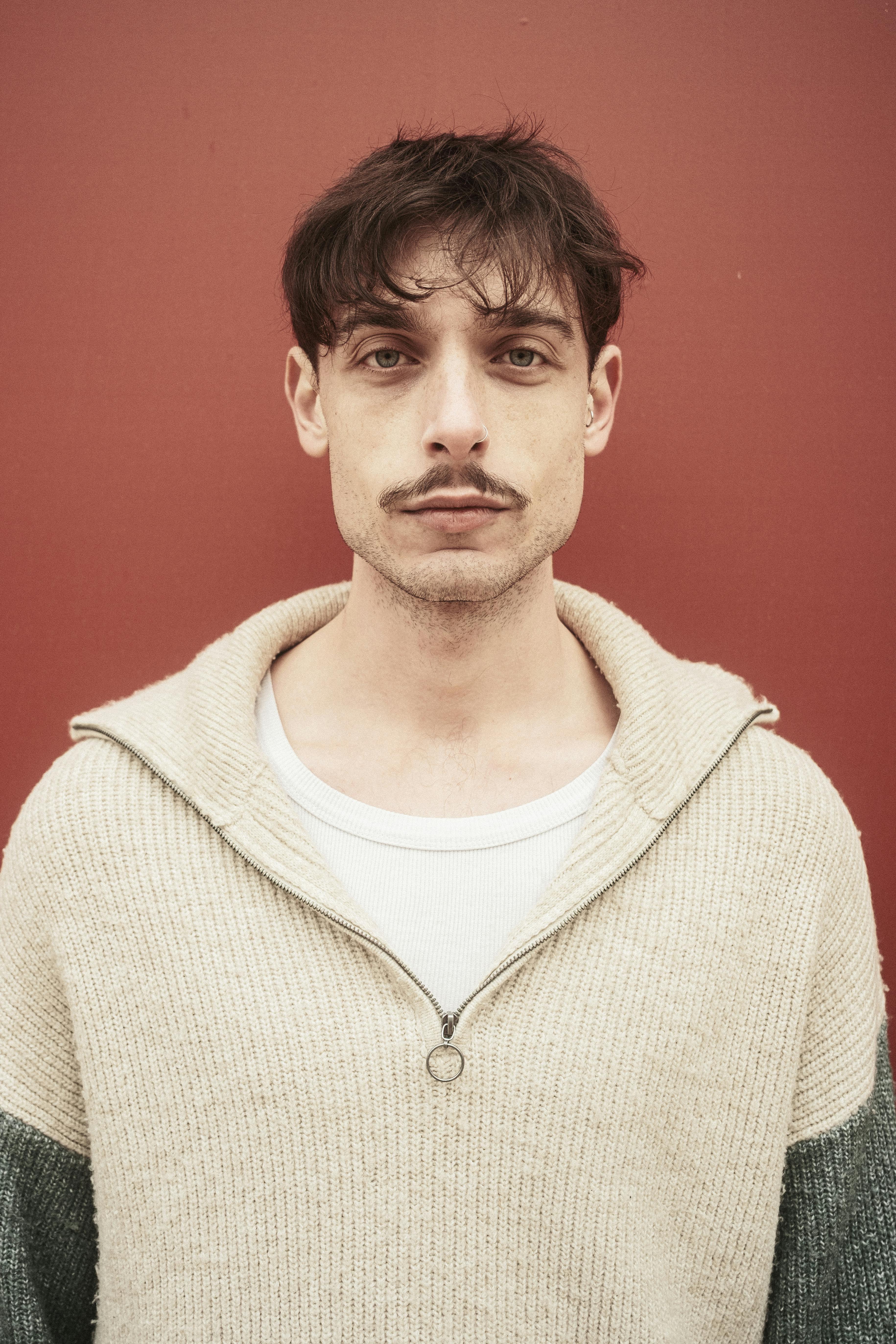 Man in a knit sweater standing against a red backdrop.