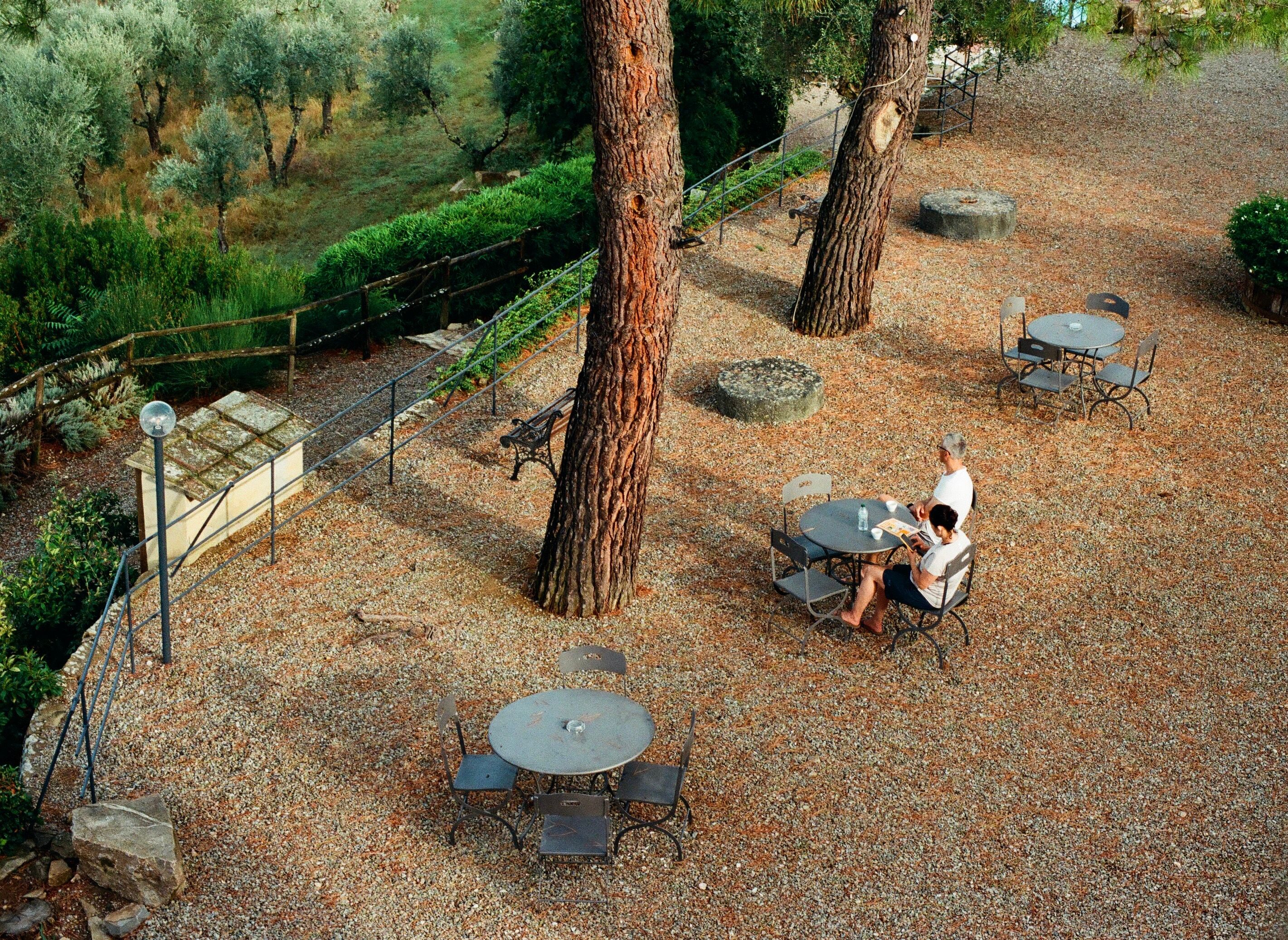 Serene outdoor café setting in Tuscany, Italy with two people relaxing under the trees on a sunny day.