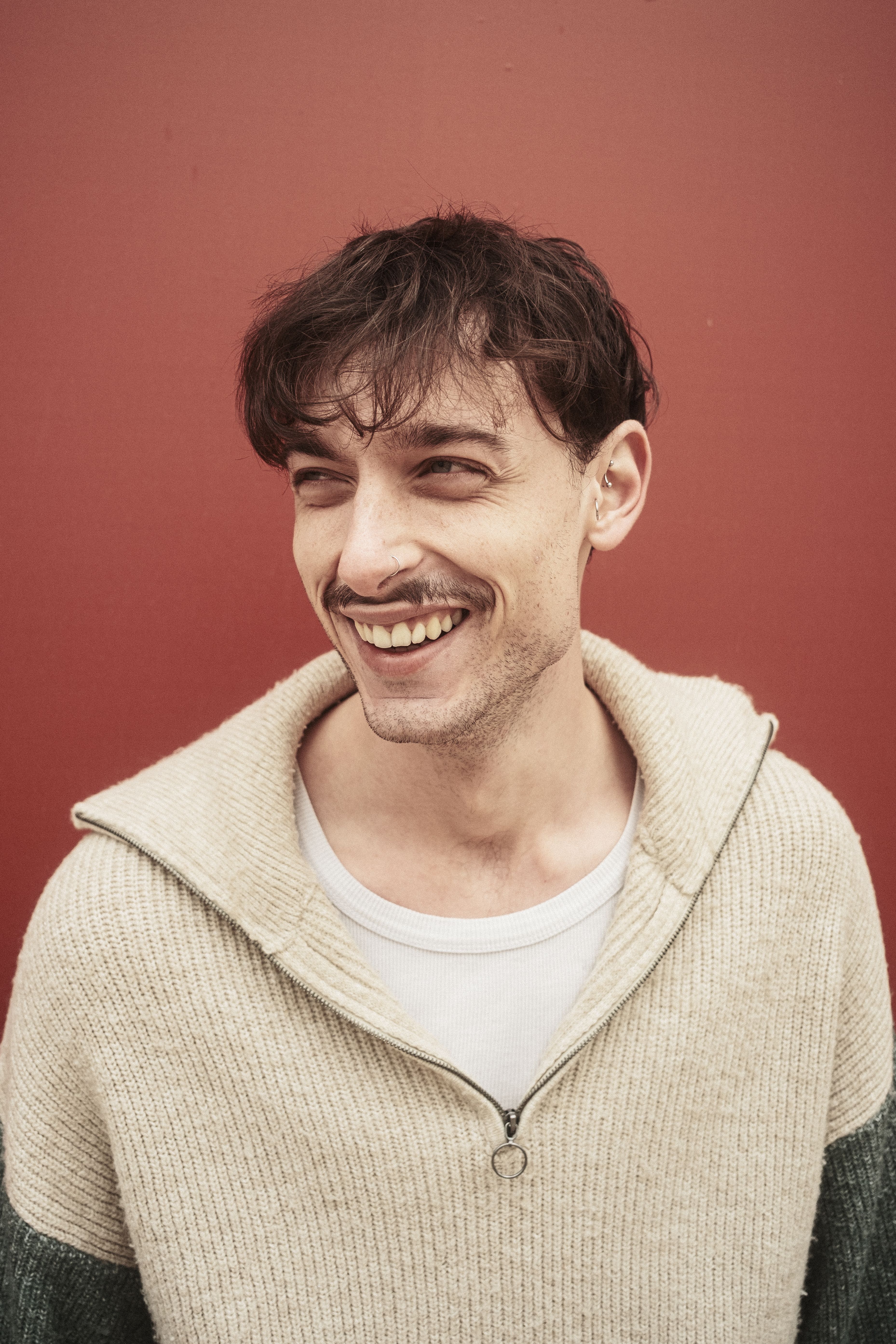 A young man smiles warmly in front of a red wall, exuding casual charm.