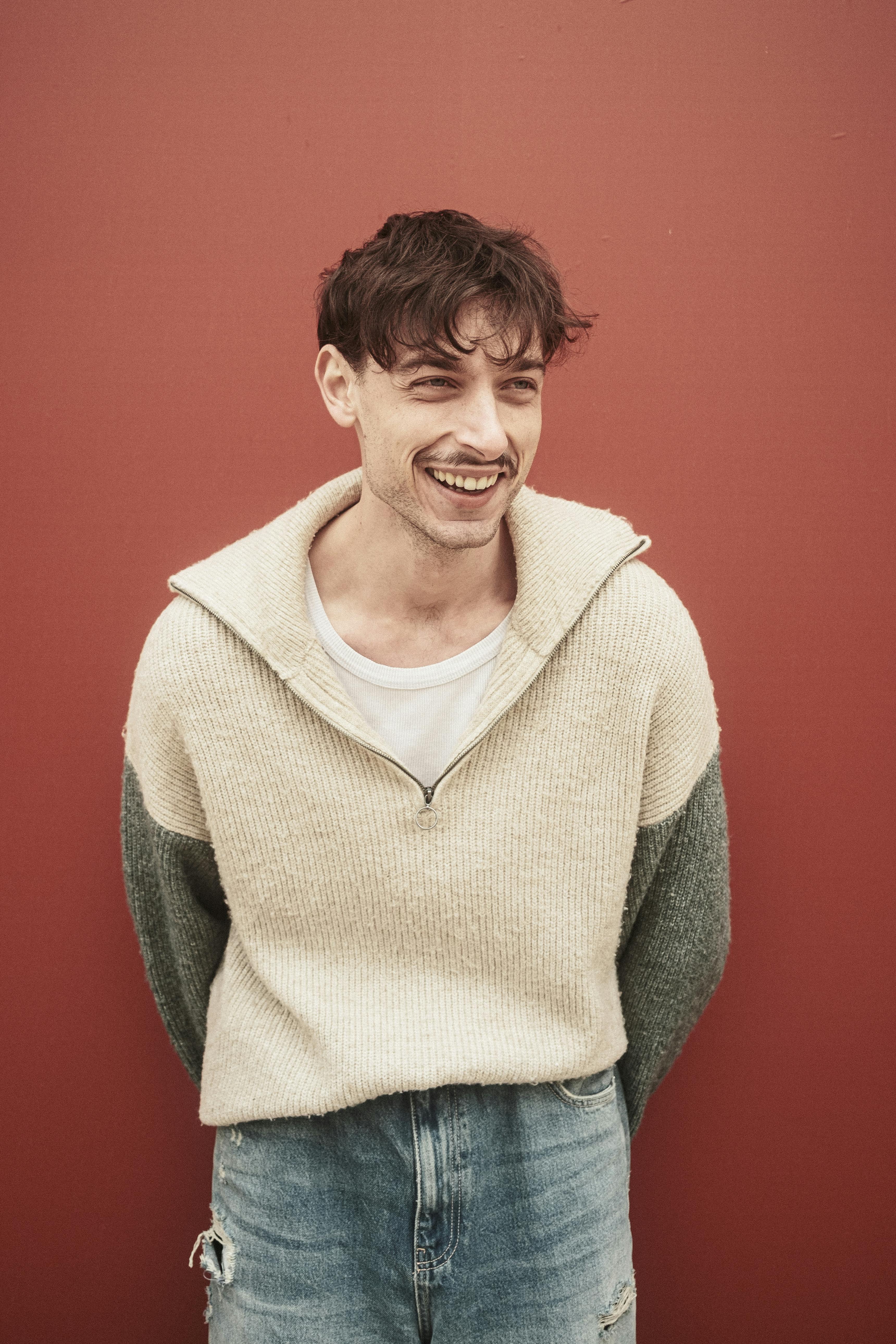 Smiling young man in casual sweater and jeans against red backdrop.