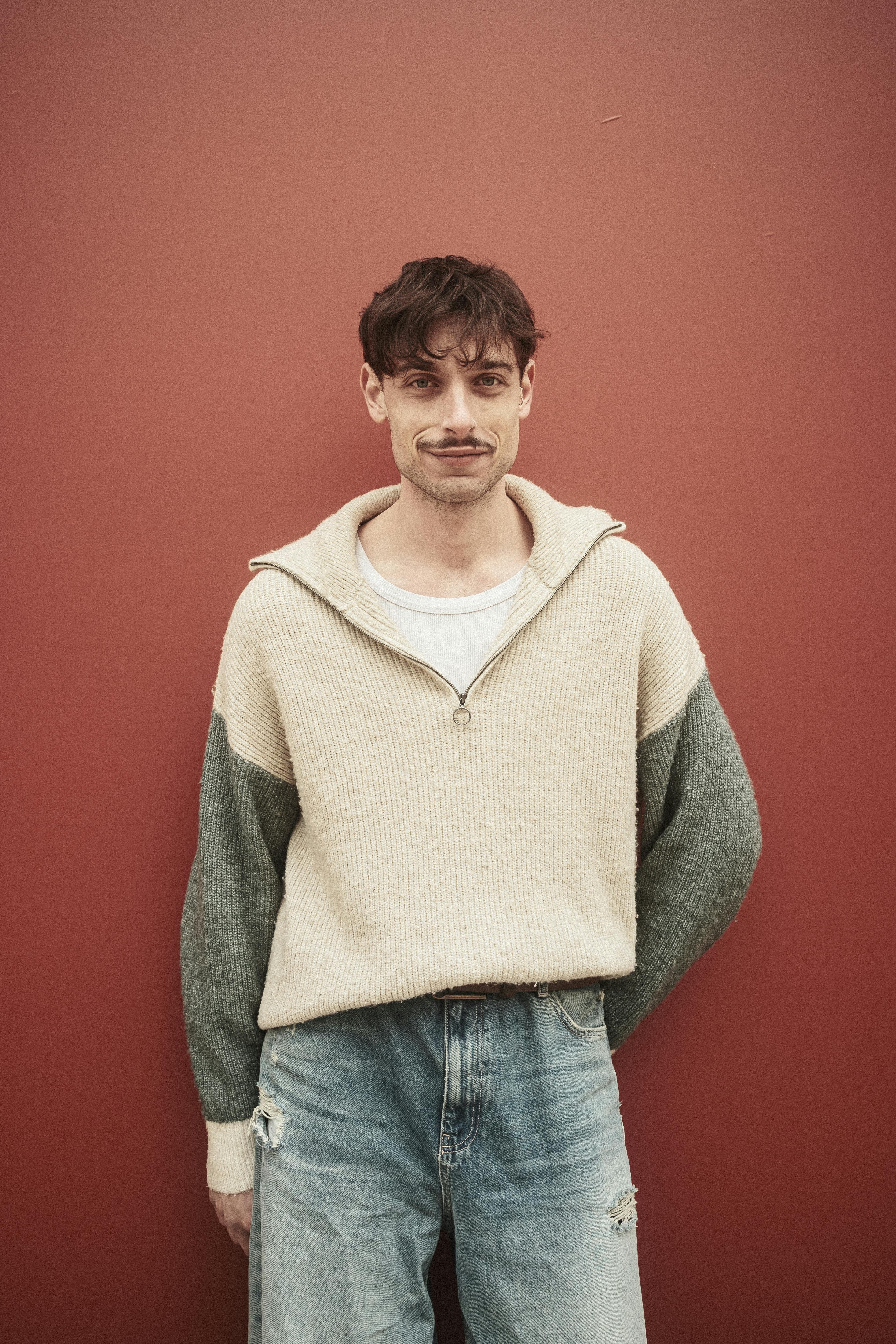 Portrait of a casually dressed man in jeans and sweater standing against a red wall.
