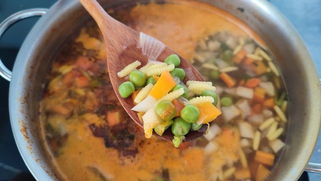 Wooden spoon lifting peas, corn and diced carrots from a simmering vegetable soup pot