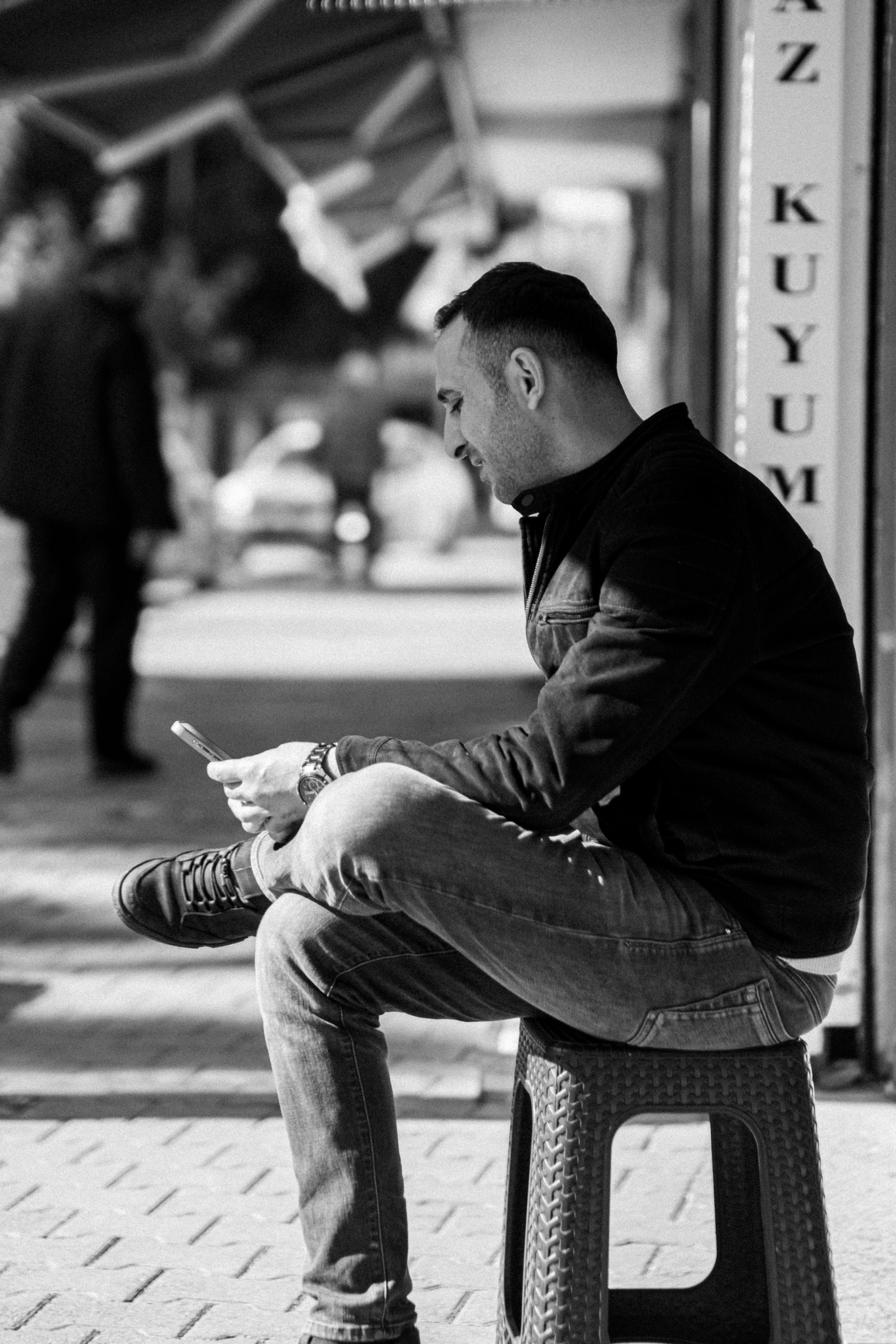 Δωρεάν Ασπρόμαυρη φωτογραφία ενός άντρα που κάθεται σε ένα σκαμπό, απορροφημένος στο smartphone του, σε εξωτερικό χώρο μια ηλιόλουστη μέρα. Φωτογραφία από στοκ φωτογραφιών