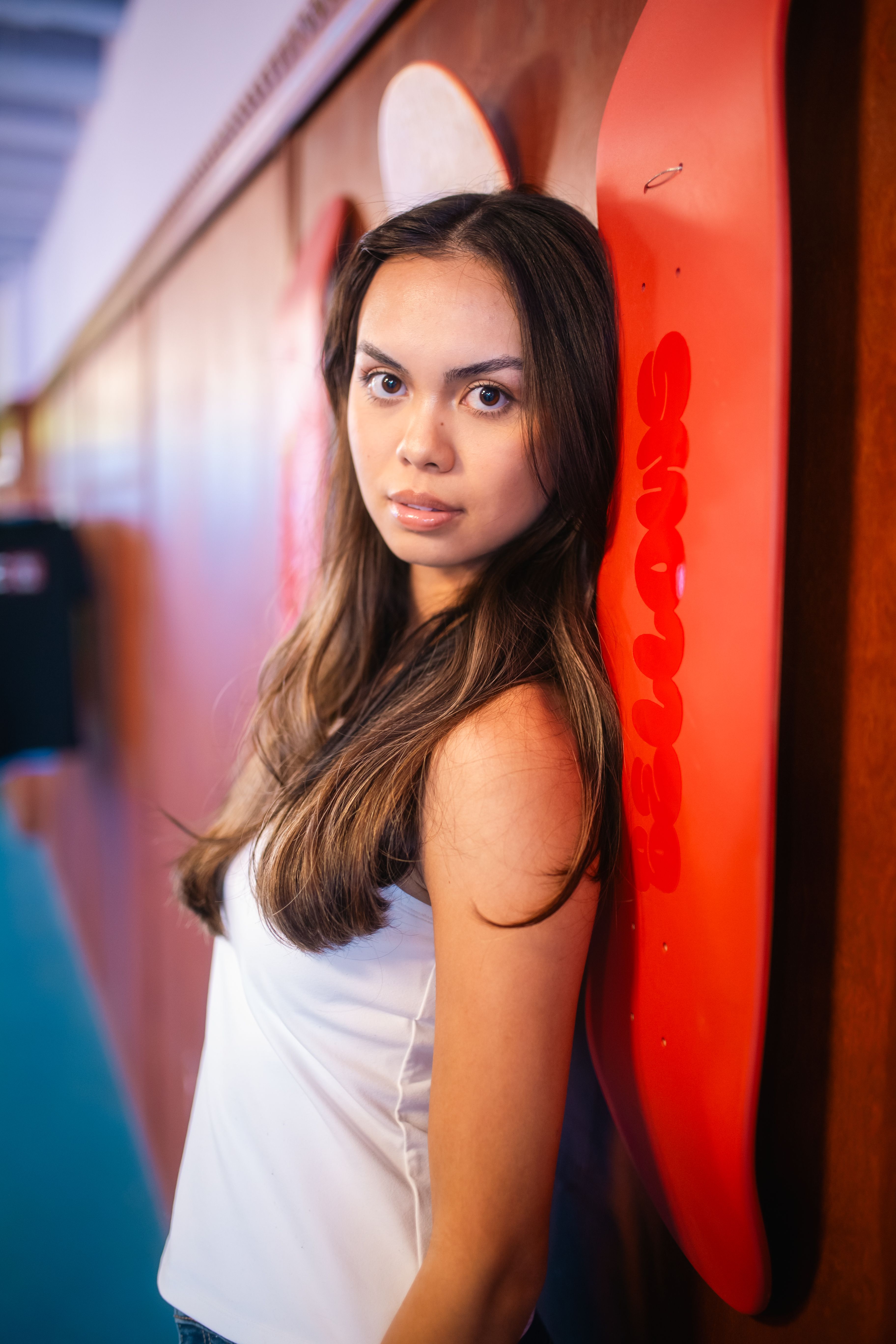 Young woman in white top posing indoors, leaning on a skateboard with vibrant colors.