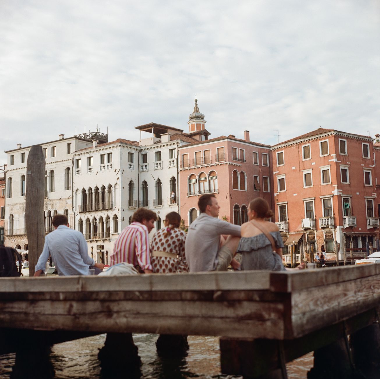 Five people sit on a dock by a canal in Venice, Italy, enjoying the view.