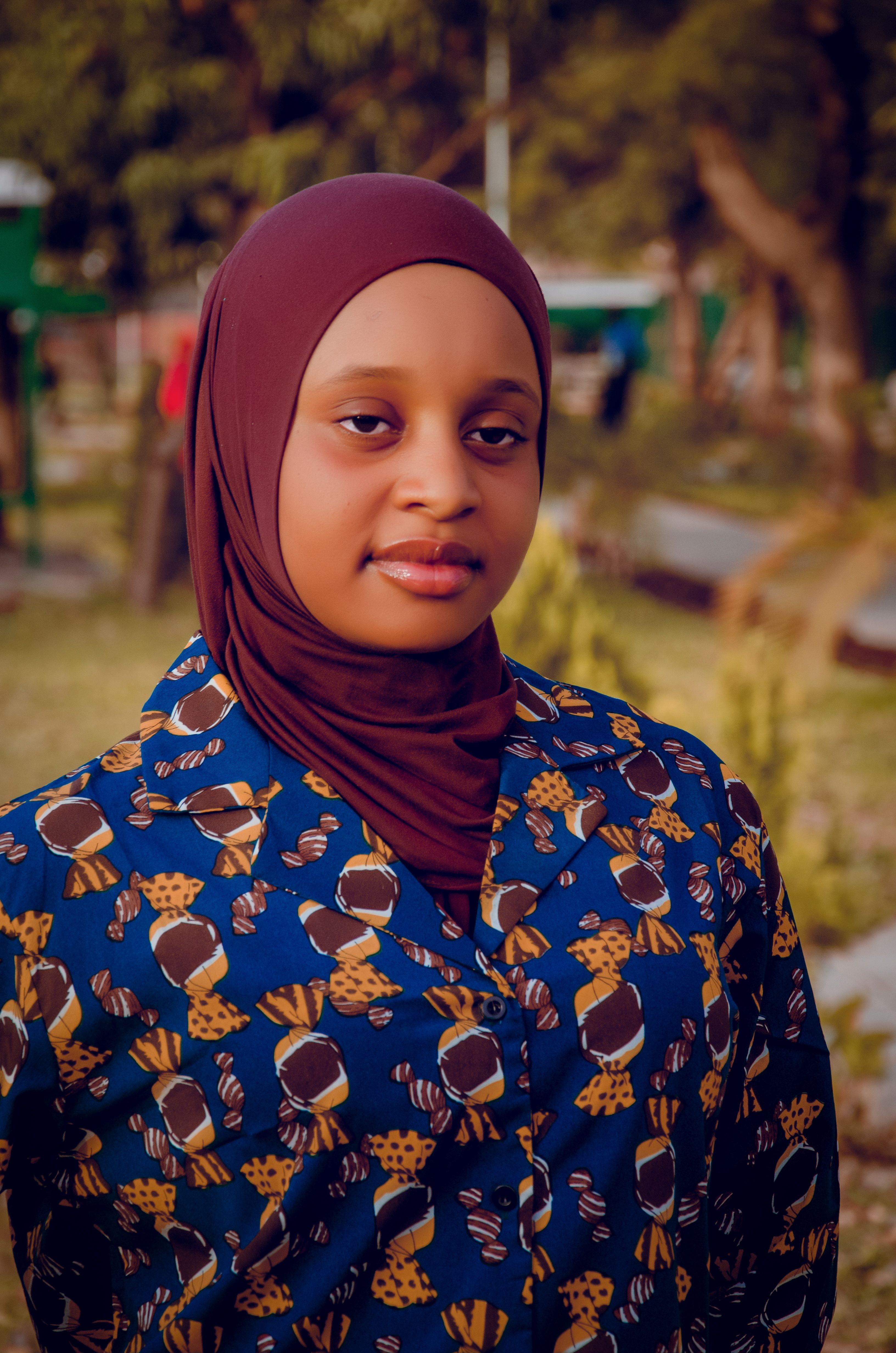 Portrait of a young woman wearing a patterned shirt and hijab in a natural outdoor setting.