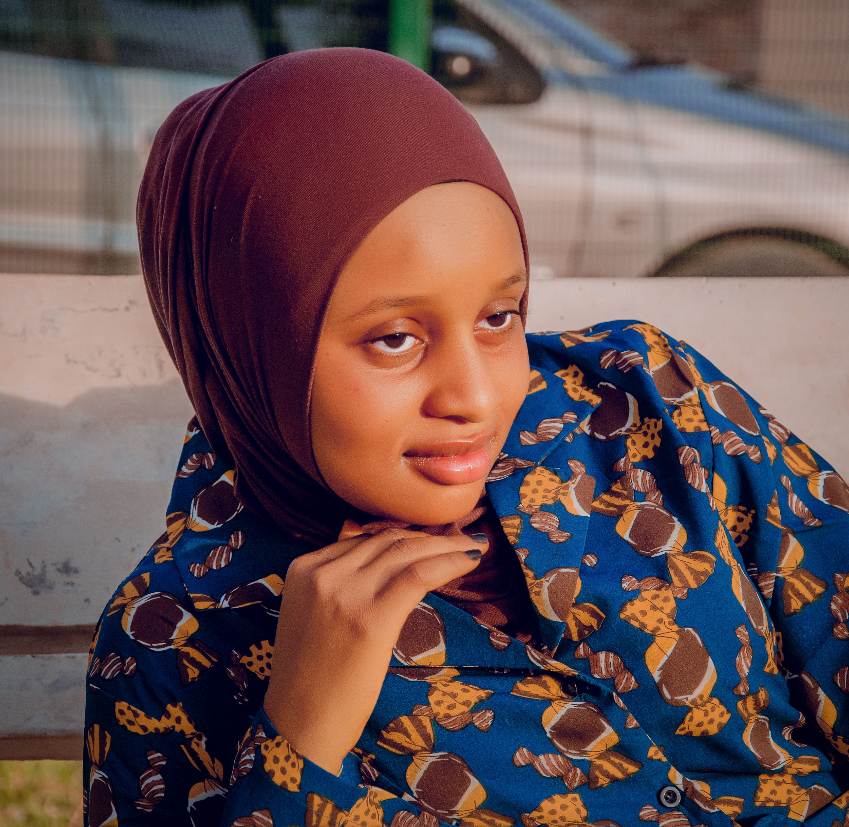 A young woman in a patterned blue shirt and hijab poses confidently outdoors.